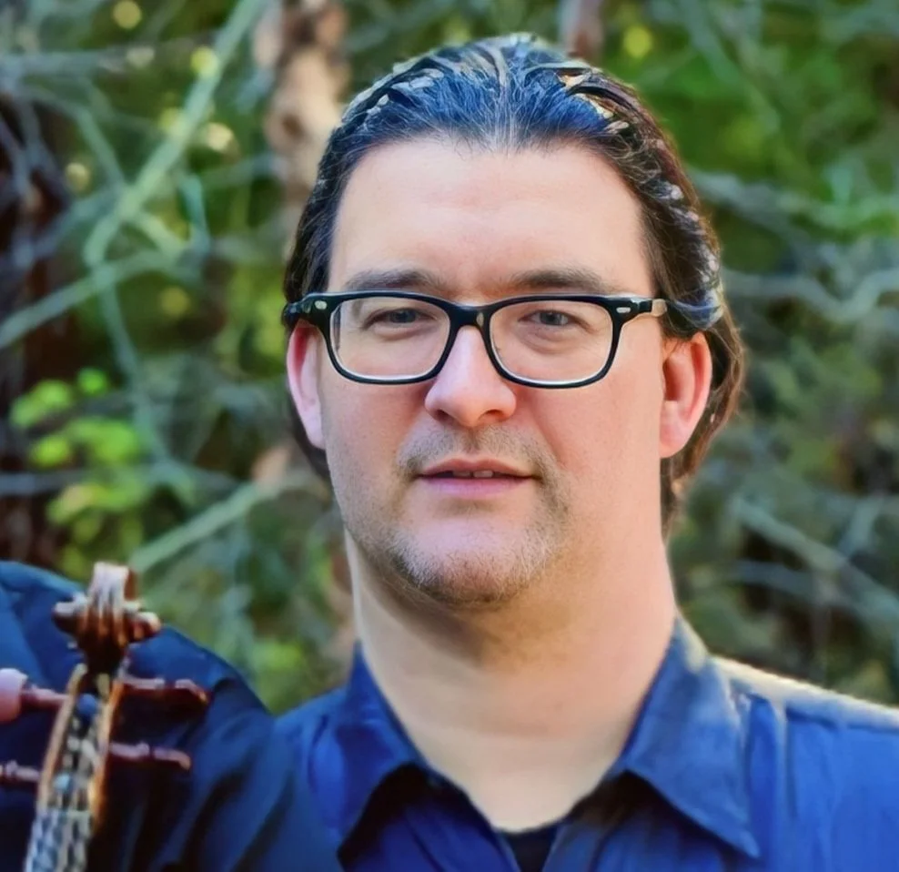 A man with glasses and dark, slightly wavy hair standing outdoors surrounded by green foliage.