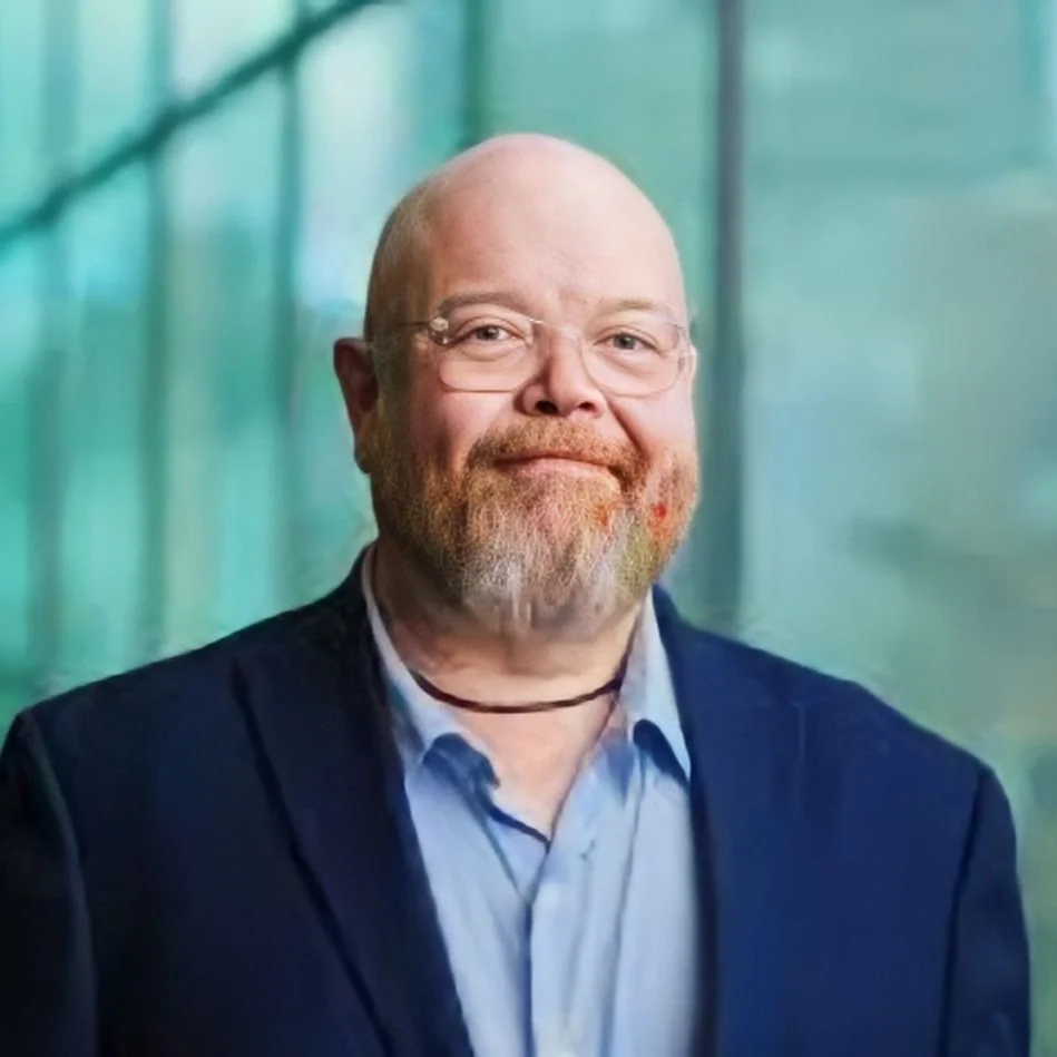 Portrait of a middle-aged man with a beard and glasses, wearing a dark blazer and a light blue shirt, standing outdoors with a blurred green background.