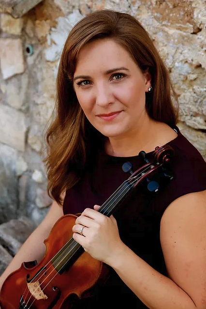 A woman with long brown hair holding a violin outdoors against a stone wall.