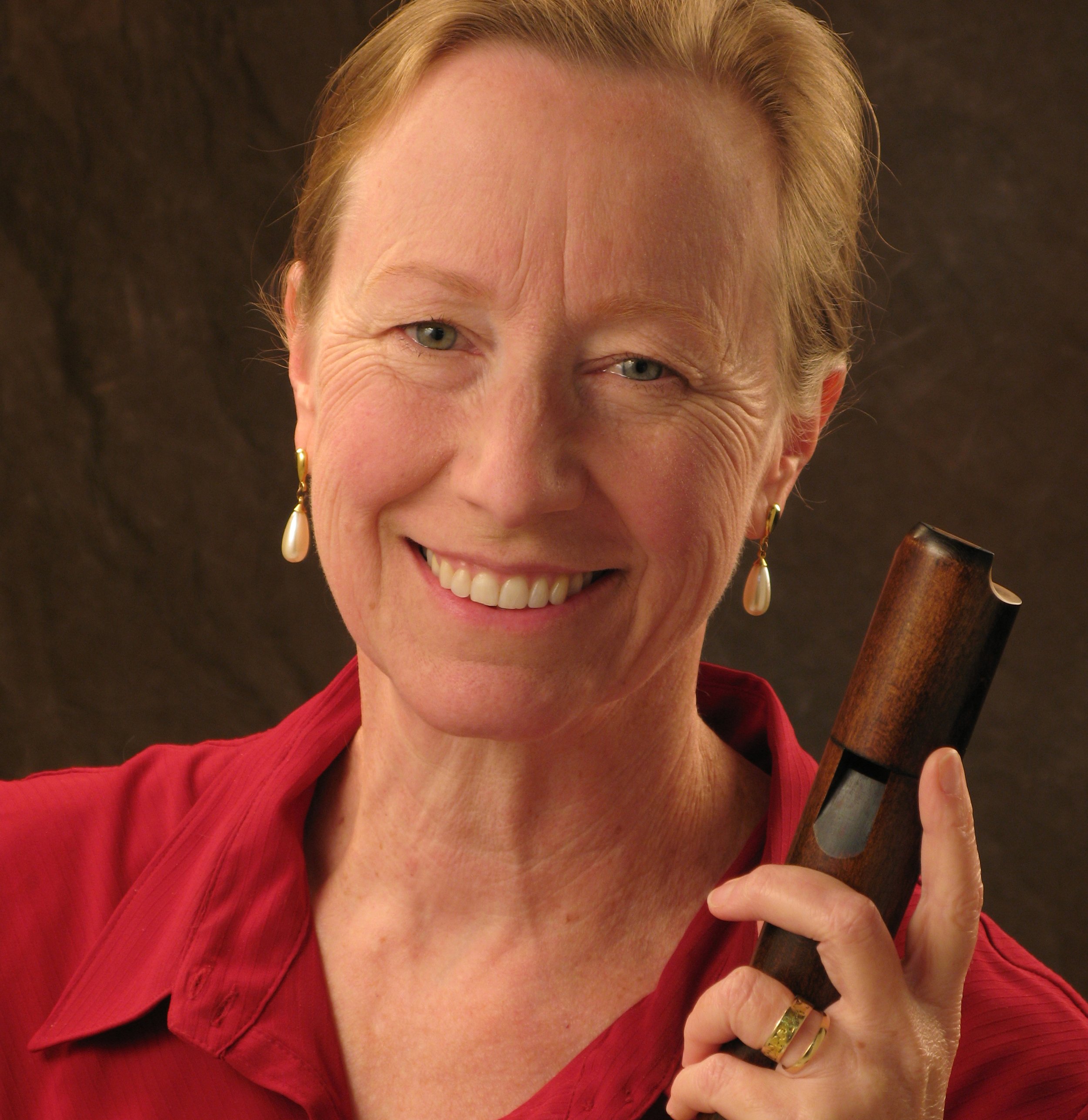 A woman with short blonde hair, smiling and holding a wooden tobacco pipe, wearing pearl earrings and a red blouse, against a dark background.