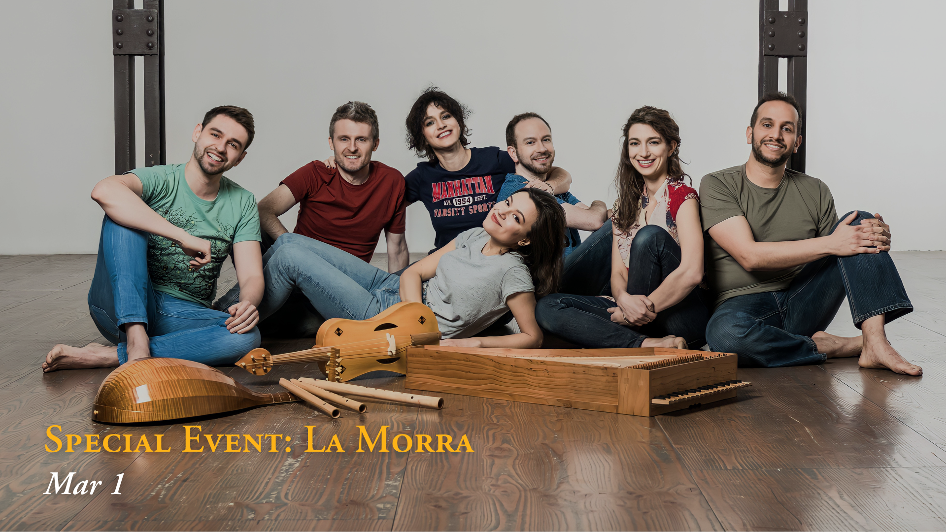 Group of seven people sitting on the floor in front of a plain wall, with musical instruments and a sign that reads "Special Event: La Morra, Mar 1."