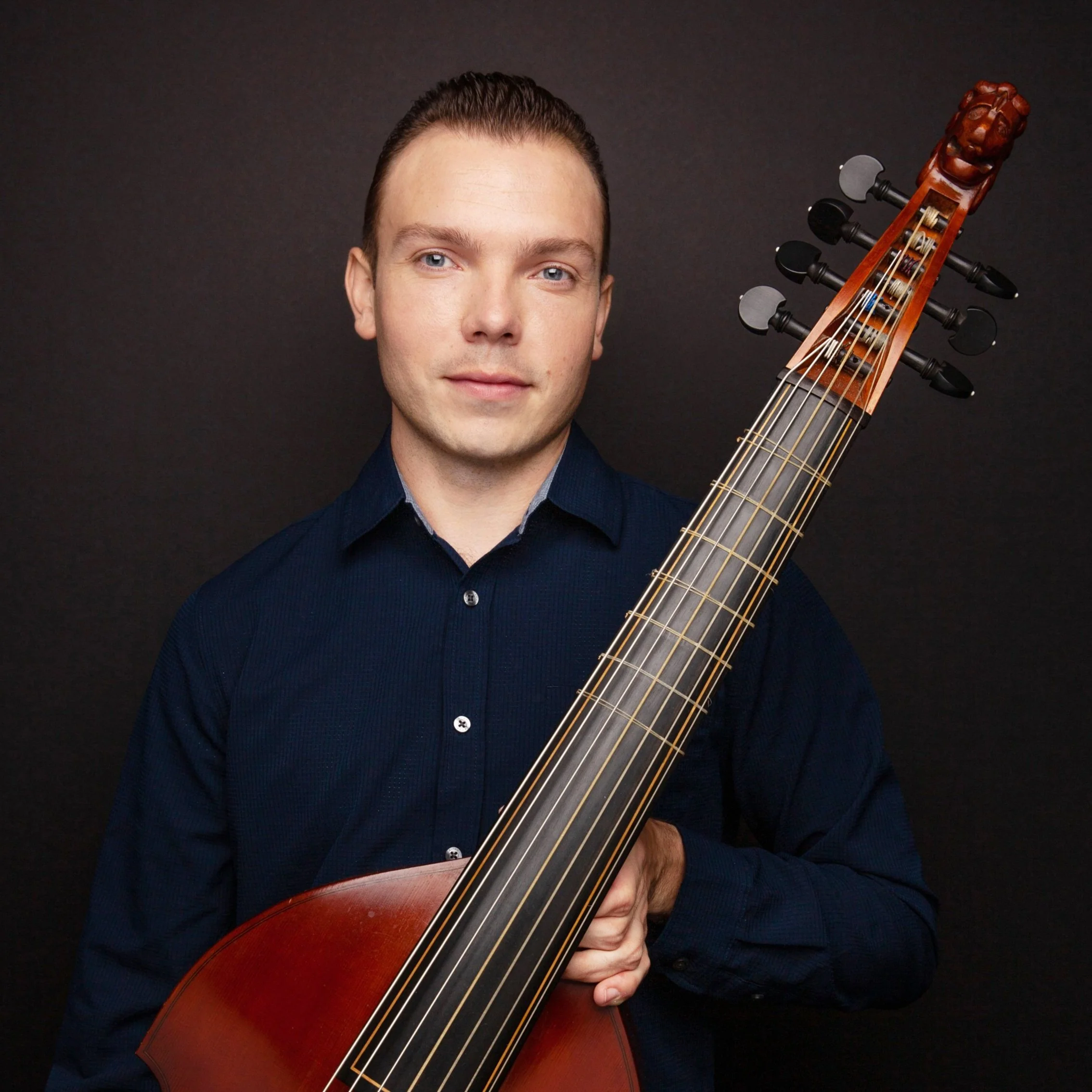A man with short dark hair and a dark blue shirt holding a large wooden stringed instrument, likely a theorbo, against a plain dark background.