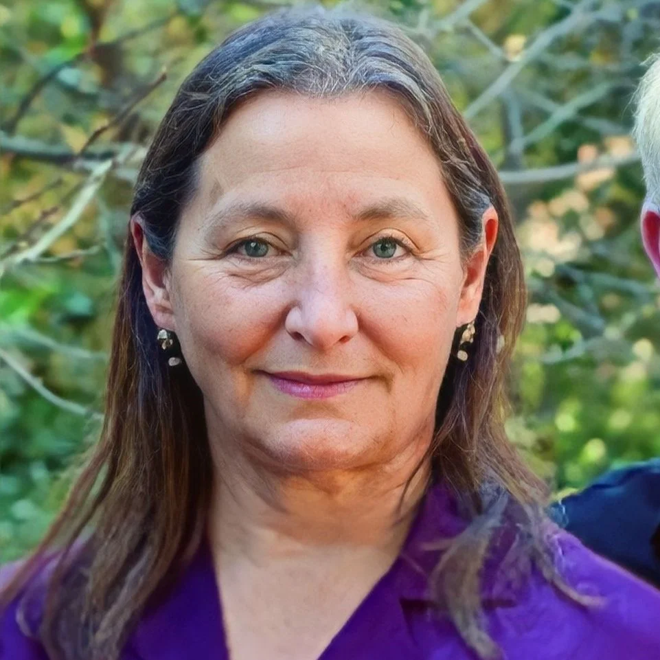 Close-up portrait of a middle-aged woman with light skin, long brown hair, and blue eyes, wearing earrings and a purple top, outdoor background with green foliage.