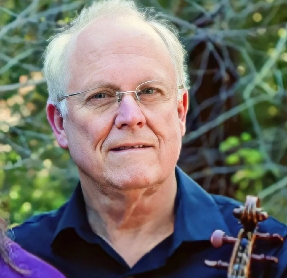 A portrait of an older man with light hair, glasses, and a slight smile, outdoors with blurry green foliage in the background.