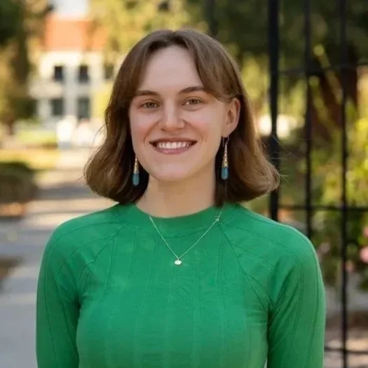 A young woman with short brown hair, smiling, wearing a green top, earrings, and a necklace, outdoors with trees and a metal gate in the background.