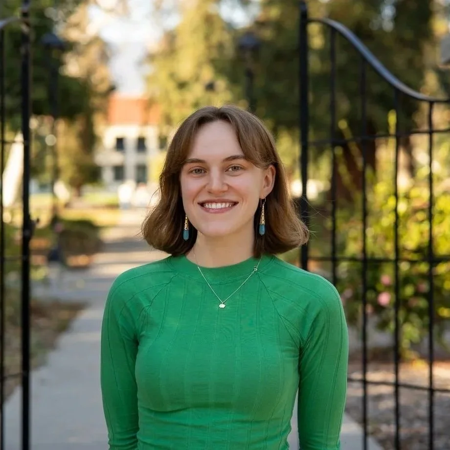 A young woman with shoulder-length brown hair, smiling, wearing a green long-sleeve top, earrings with a blue design, and a necklace with a small pendant. She is outdoors near a black metal fence, with trees and a building in the background.