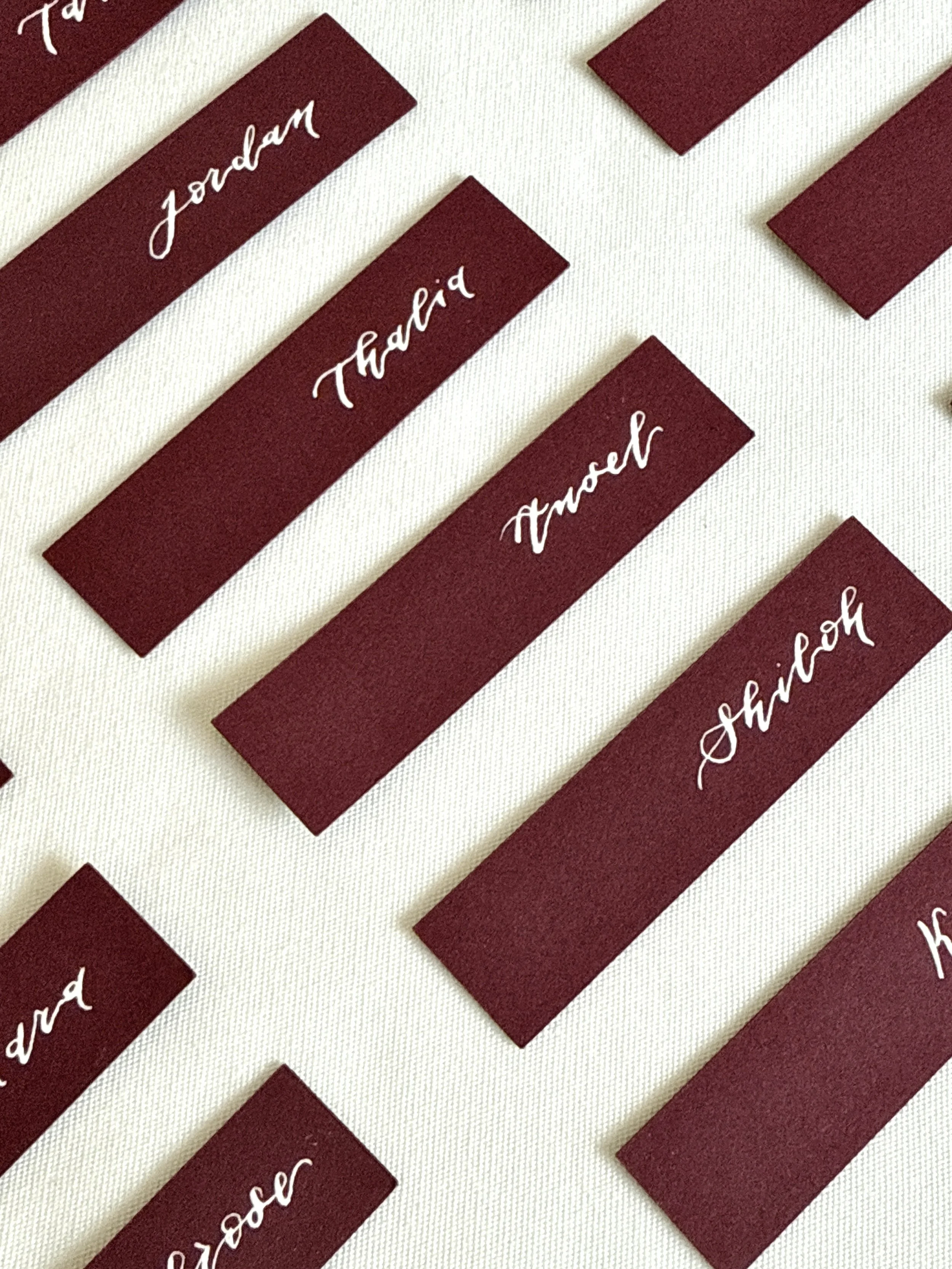 Maroon wedding place cards with white calligraphy writing for different individuals, including Jordan, Thalia, Traci, and Shahad, on a white fabric background.