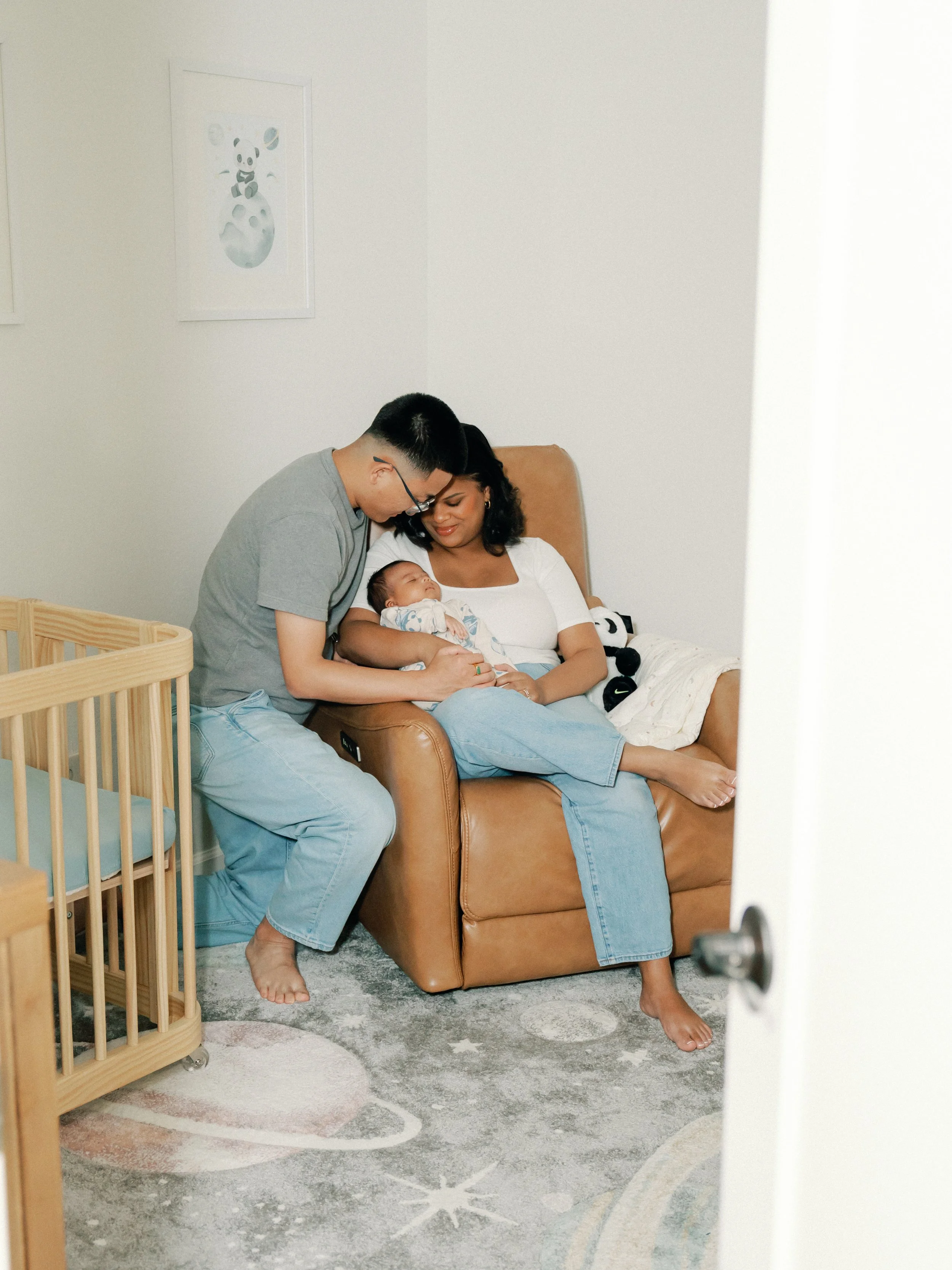 A family of three with a newborn sitting on a tan armchair in a nursery. The father, wearing glasses and a gray t-shirt, leans over to look at the baby. The mother, dressed in white, holds the baby on her lap. A wooden crib is visible to the left, and framed artwork with cartoon bears hangs on the wall behind them.