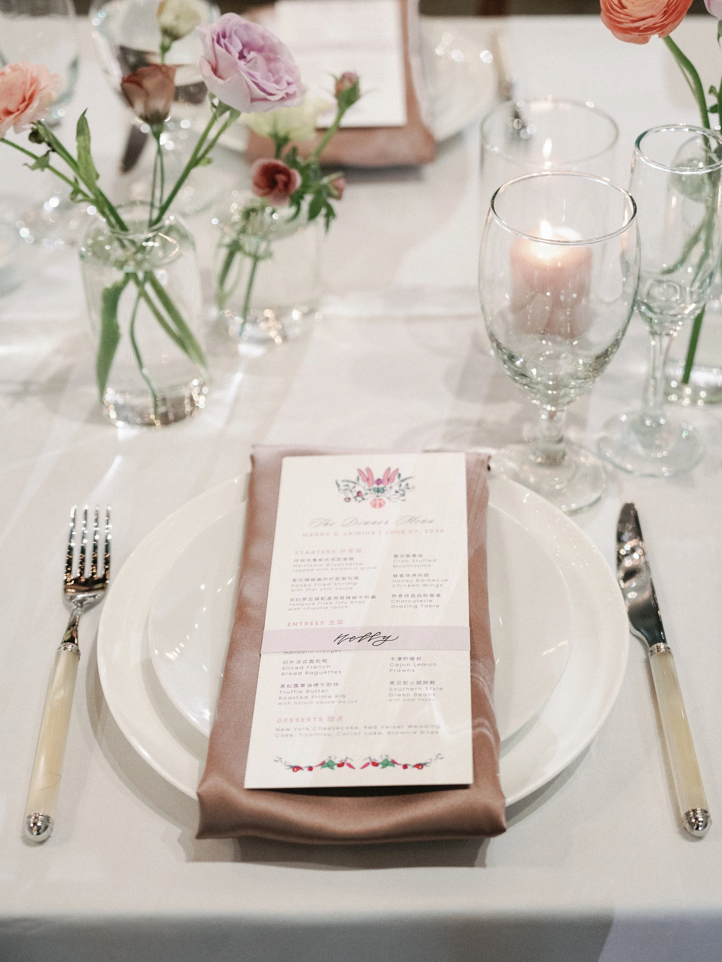 A place setting at a formal event with a white plate, silver cutlery, a pink napkin, a decorated menu, floral centerpieces, and a lit candle in a glass holder.
