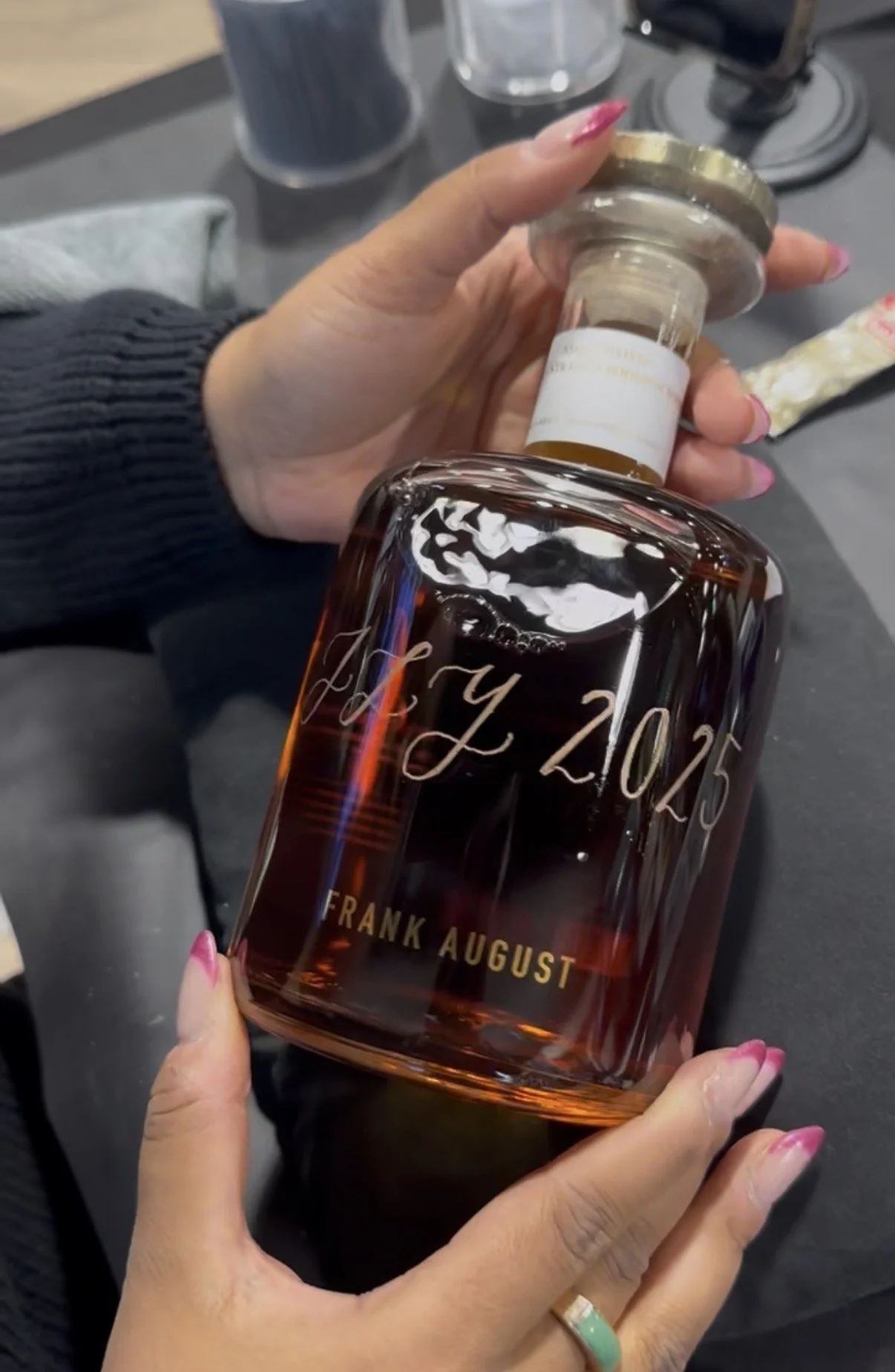 A person holding a whiskey nottle with an engraved inscription. The whiskey decanter has the name 'Frank August' and a date 'JLY 2025' engraved on it.