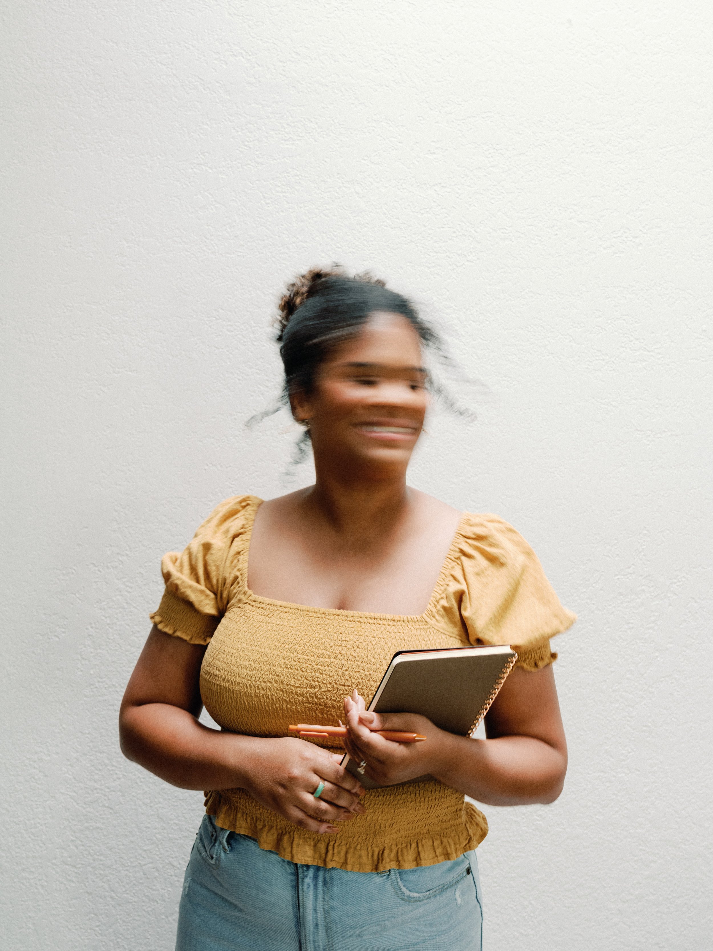 A Black woman with dark hair tied in a bun, wearing a yellow top with puffed sleeves, stands against a textured white wall, holding a notebook and orange pen, smiling.