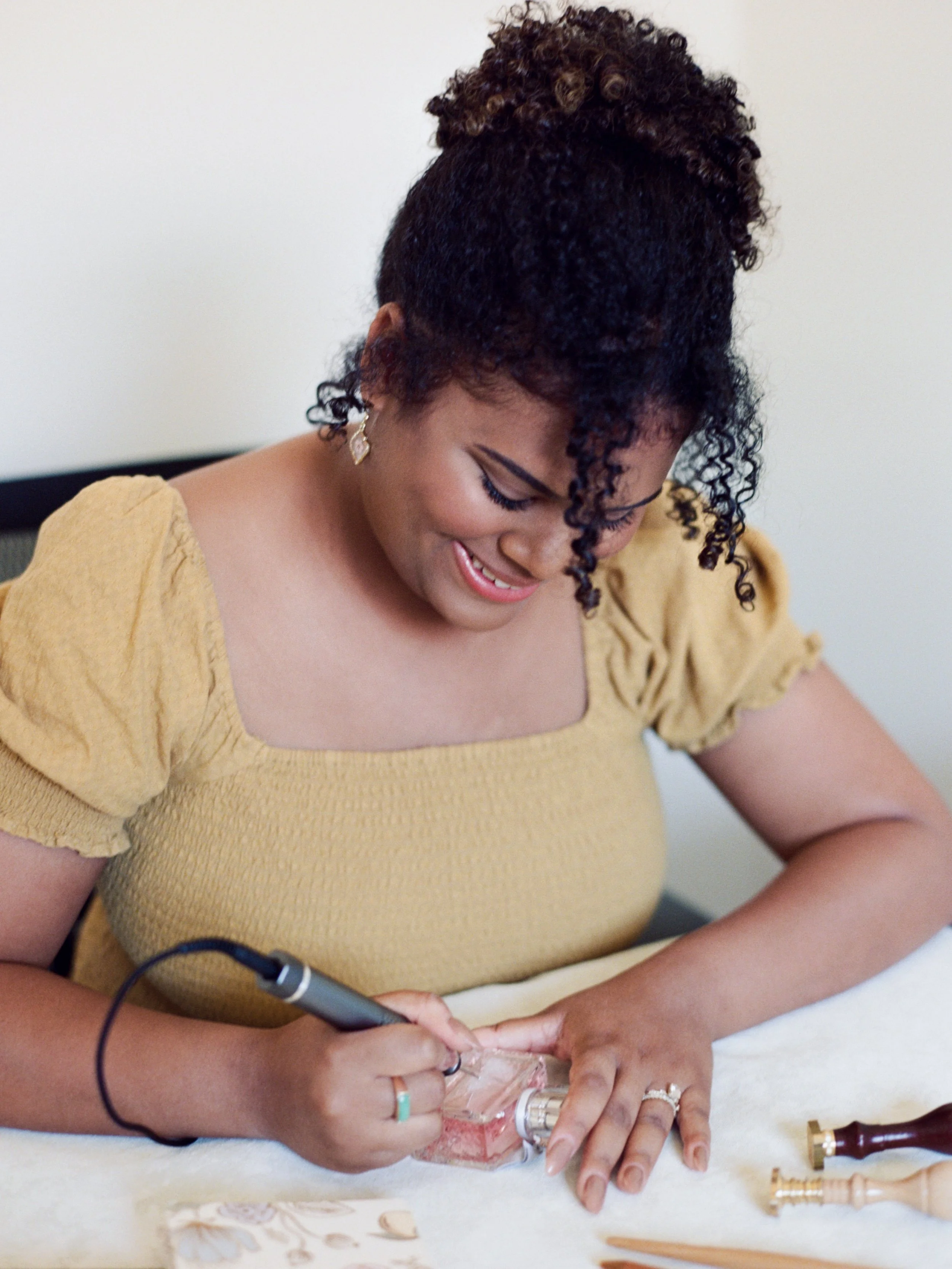 Black woman with curly hair in a bun, wearing a yellow top, using a tool to engrave a perfume bottle.