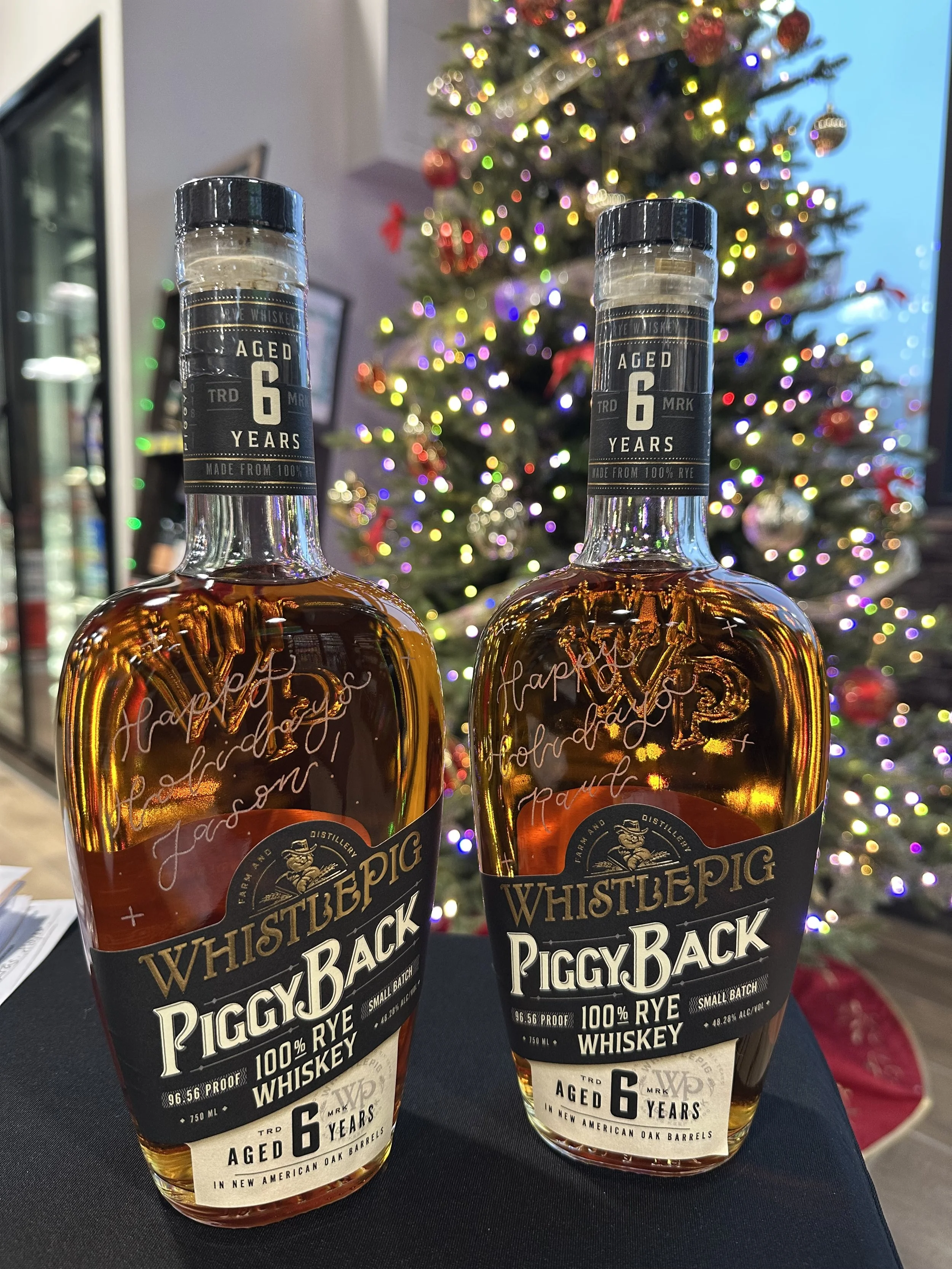 Two hand engraved bottles of WhistlePig PiggyBack whiskey are placed on a surface with a decorated Christmas tree in the background.