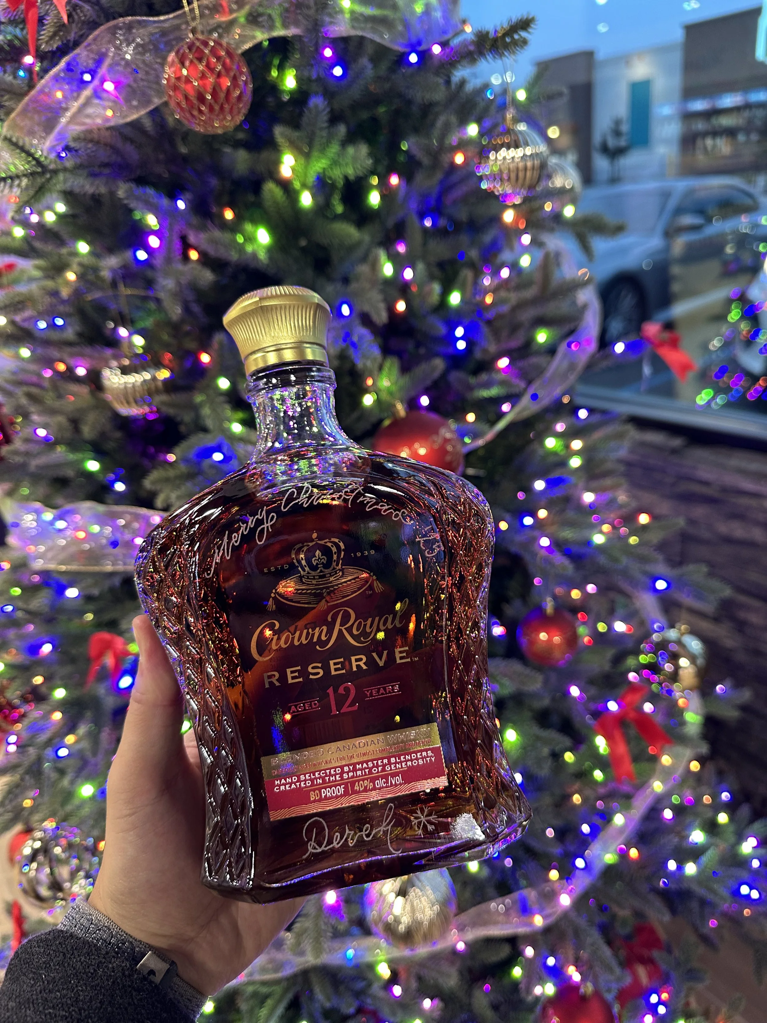 Hand holding a hand engraved Crown Royal Reserve 12-year whiskey bottle in front of a decorated Christmas tree with colorful lights and ornaments.