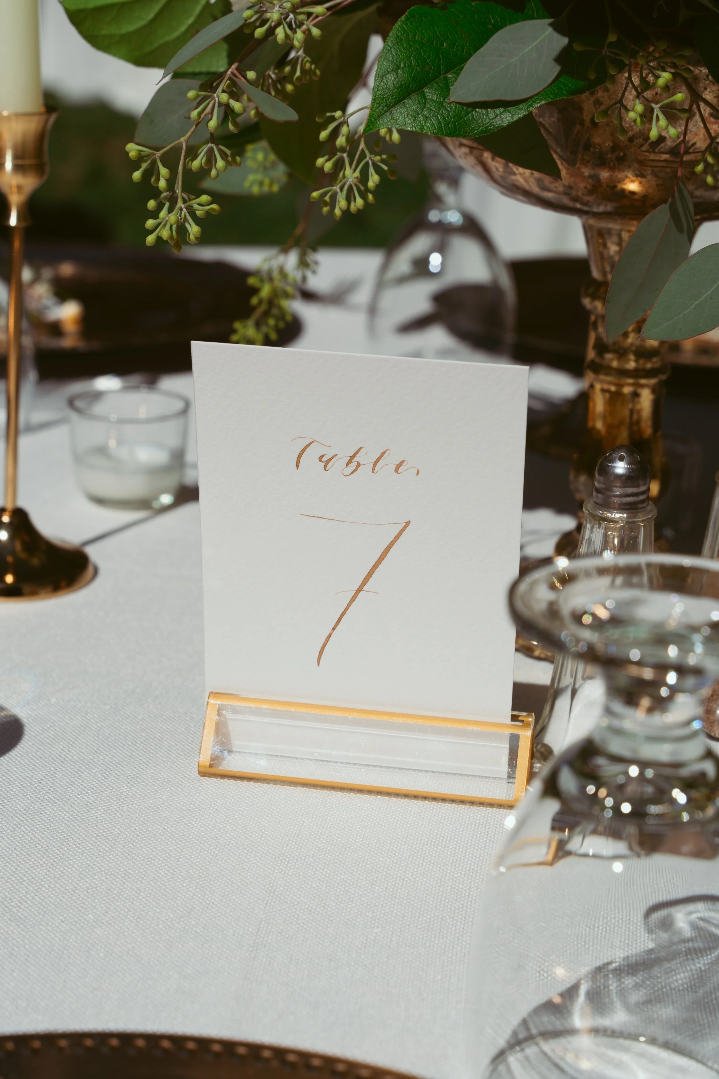 A table card with 'Table 7' written on it, placed on a white tablecloth, surrounded by glassware, candles, and a floral centerpiece with green leaves and small buds.