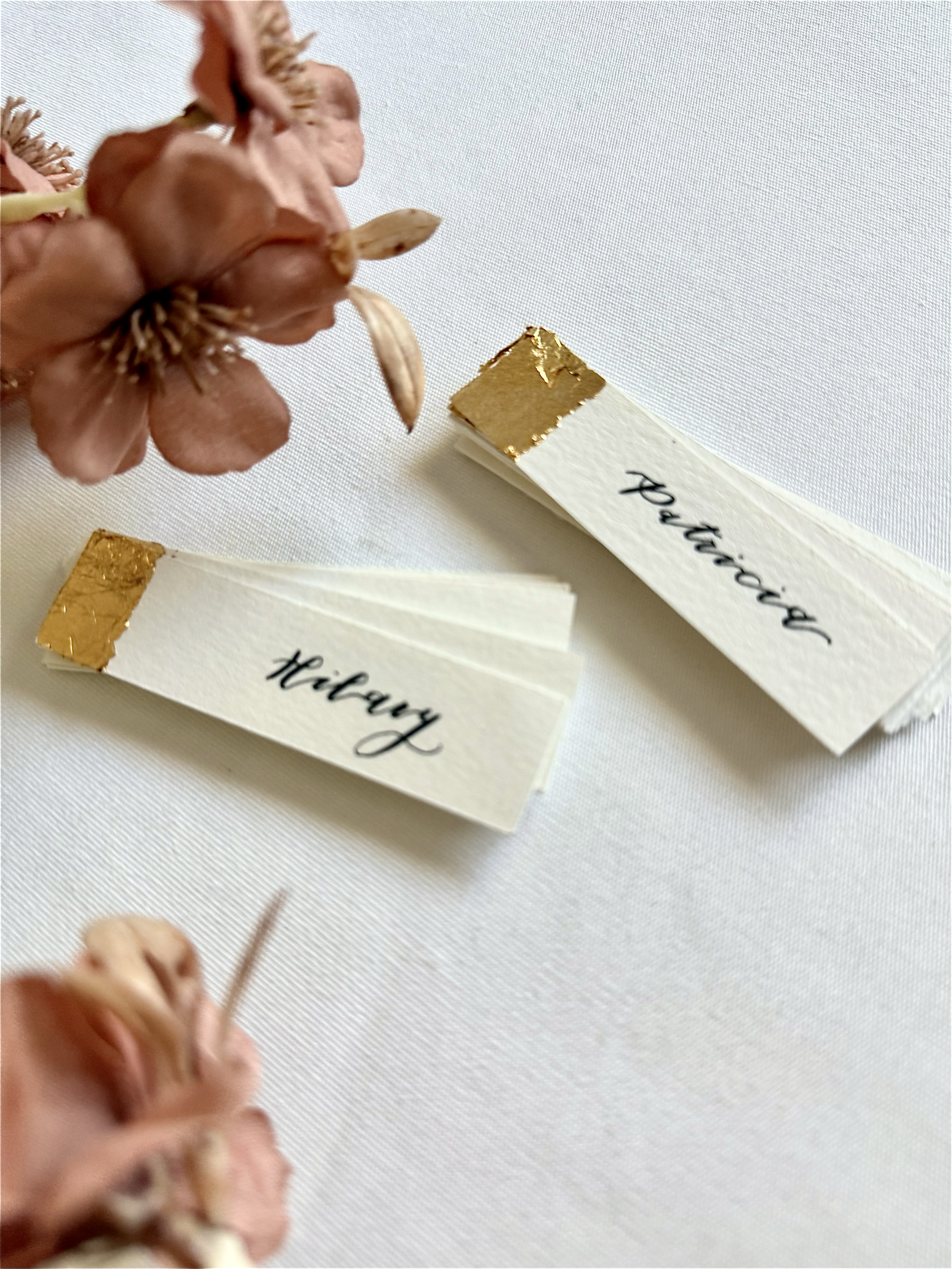 Wedding place cards labeled "Hilary" and "Patricia" with gold accents, next to a dried flower on a white surface.