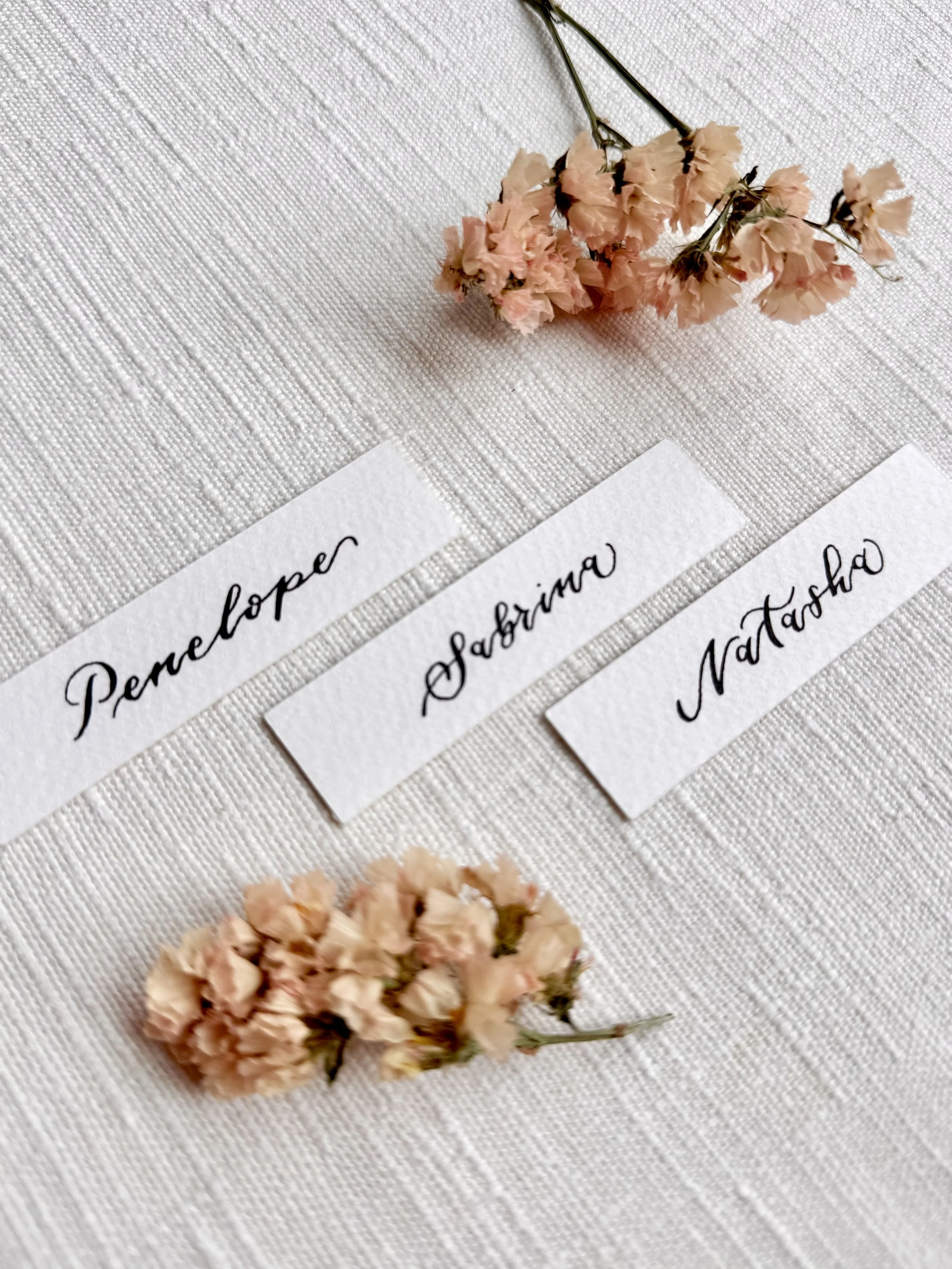Three wedding name cards or wedding place cards reading "Penelope," "Sabrina," and "Natasha" arranged on a white textured fabric, with pink dried flowers above and below the tags.