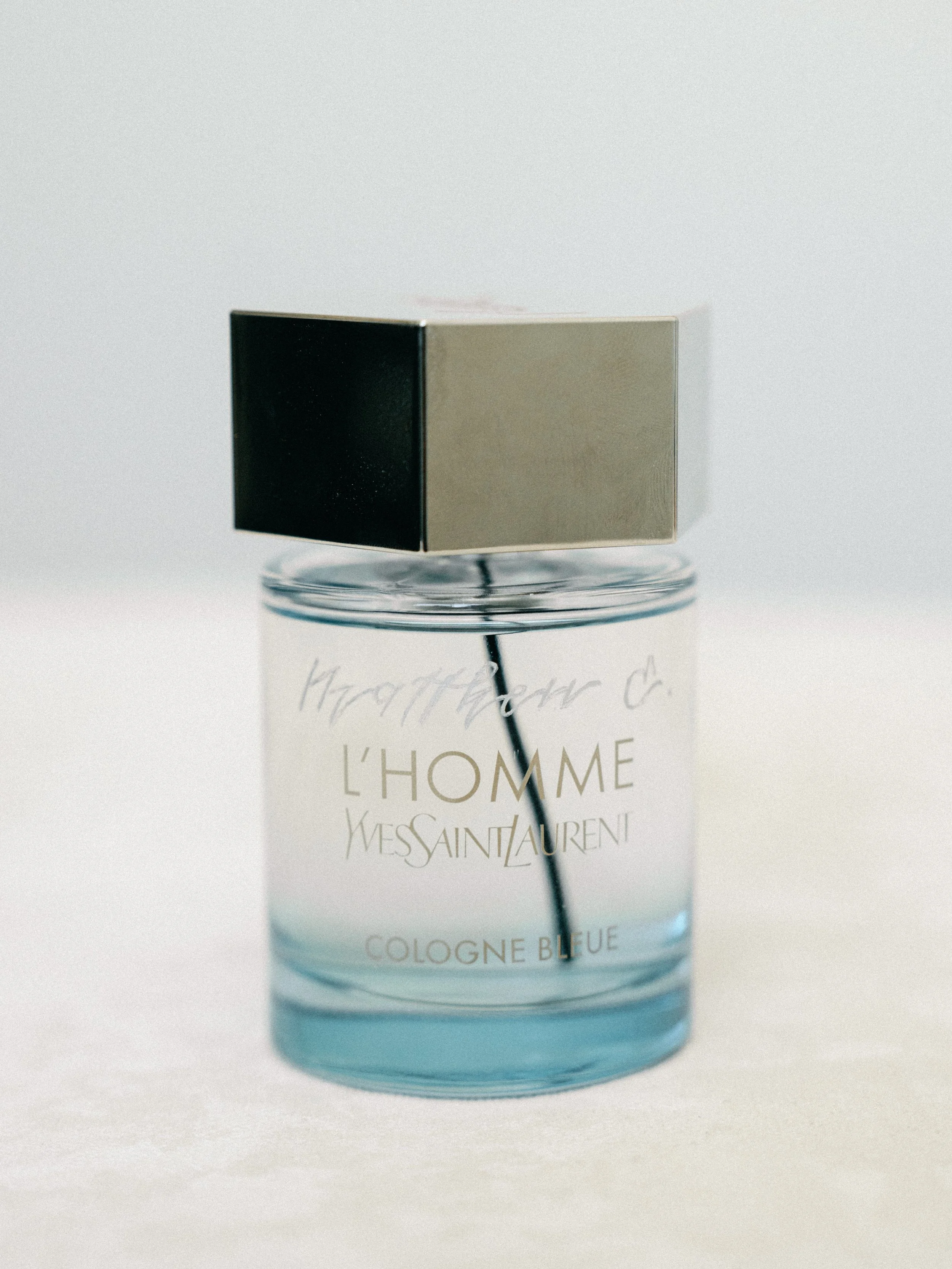 An engraved bottle of Yves Saint Laurent L'Homme Cologne Bleue perfume with a silver cap, placed on a clear glass stand against a white background.