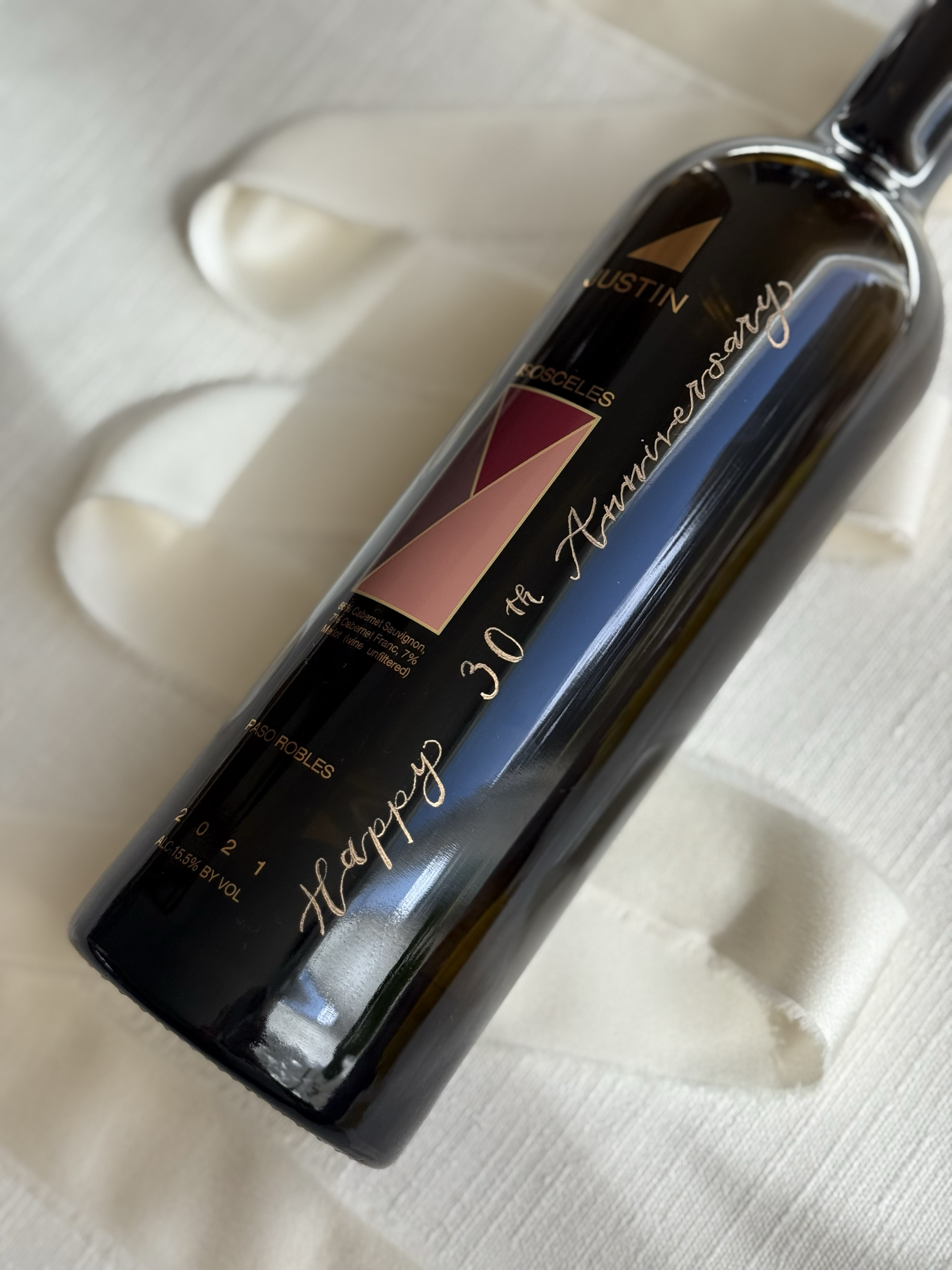 A black wine bottle with personalized engraving and gold filling and a pink label, resting on a white textured fabric with ribbon-like adornments.