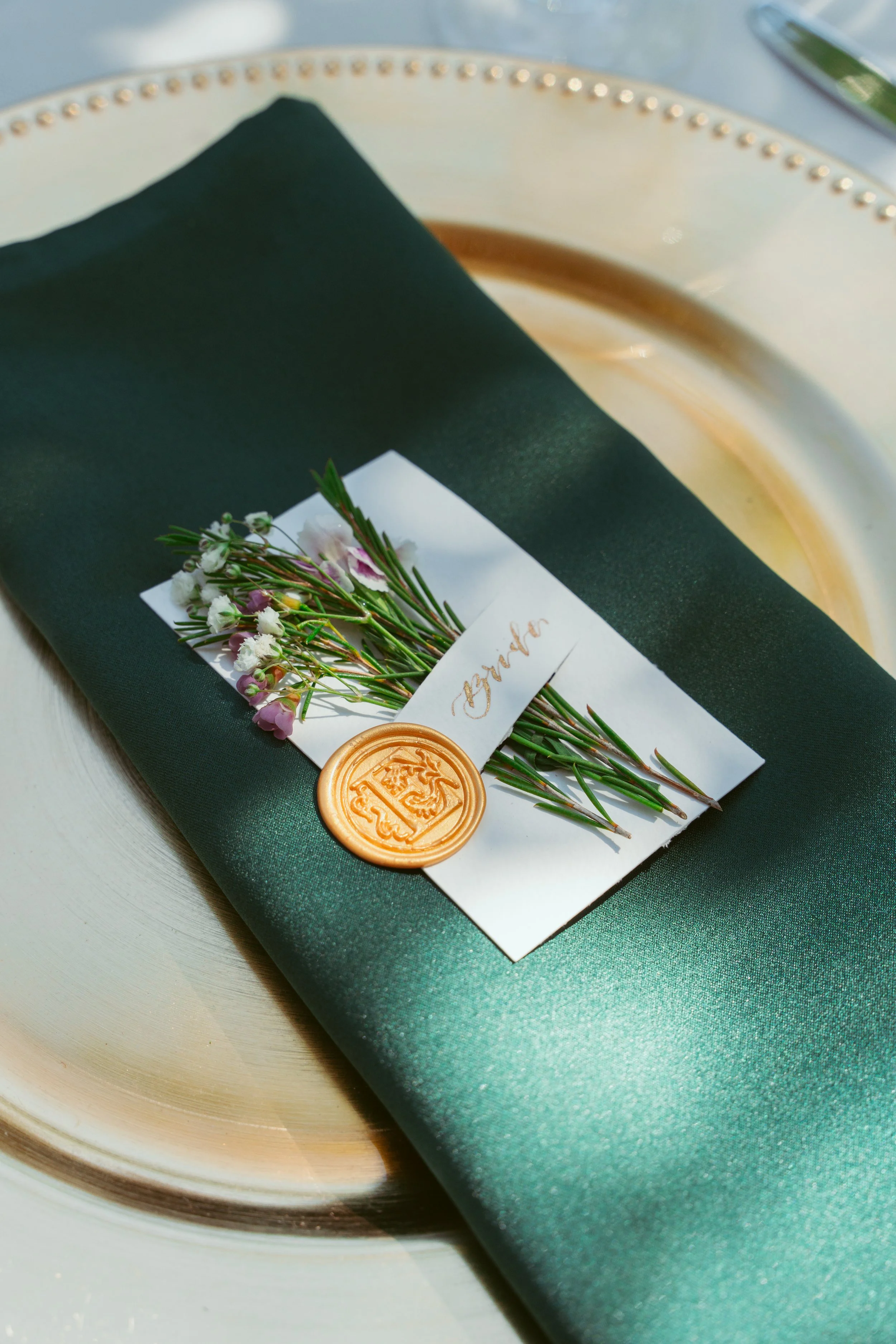 A decorative wedding place card featuring a dark green napkin, a small bouquet of white and purple flowers, a white card with a handwritten name, and a gold wax seal, placed on a cream-colored plate.