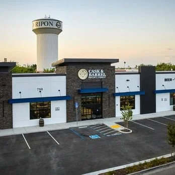 Exterior of Cask & Barrel in Ripon, California with a parking lot in front, and a red and white water tower in the background.