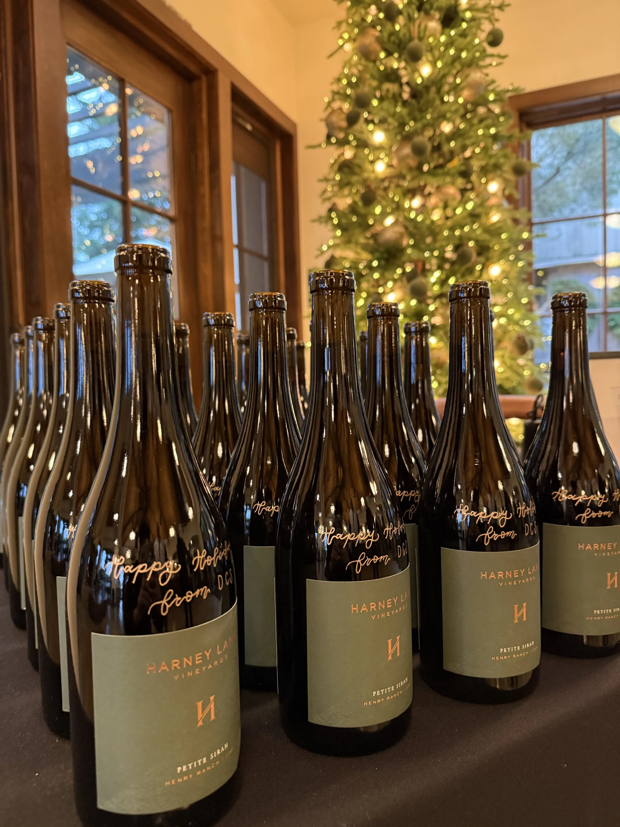 Multiple bottles of hand engraved Harney Lane Vineyard Petite Sirah wine bottles on a table in front of a decorated Christmas tree with lights in a room with wooden window frames.