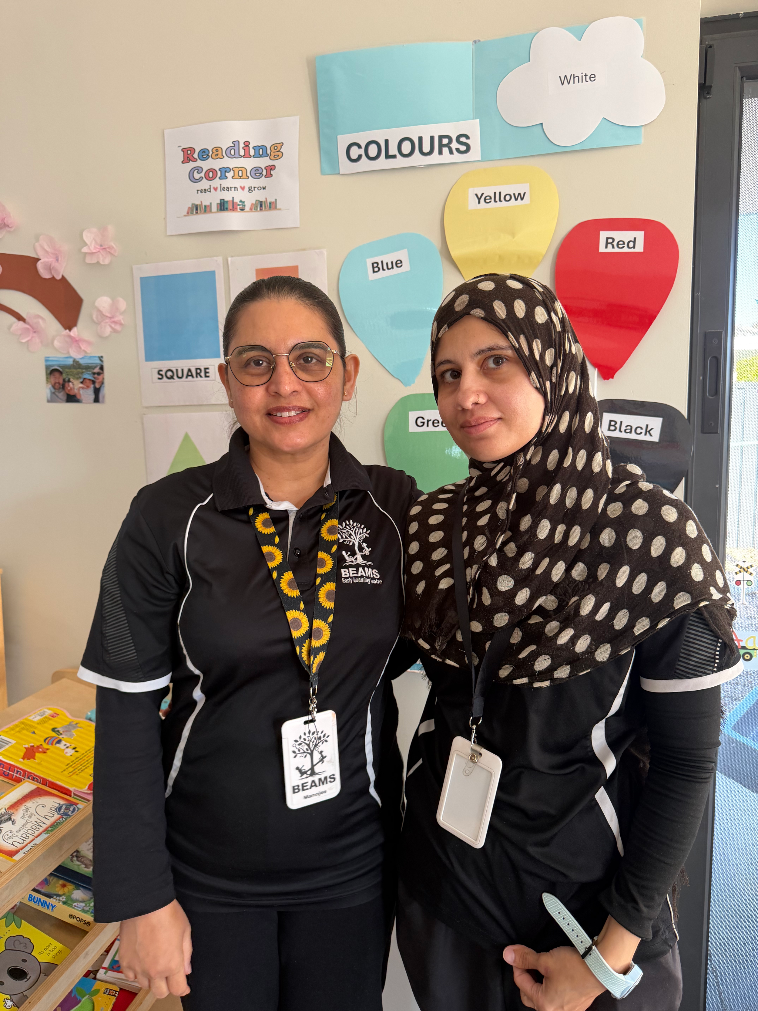 Two women workign at Beams wearing black Beams uniforms with name tags stand in front of a wall displaying children's artwork, including paintings with stars and red shapes.