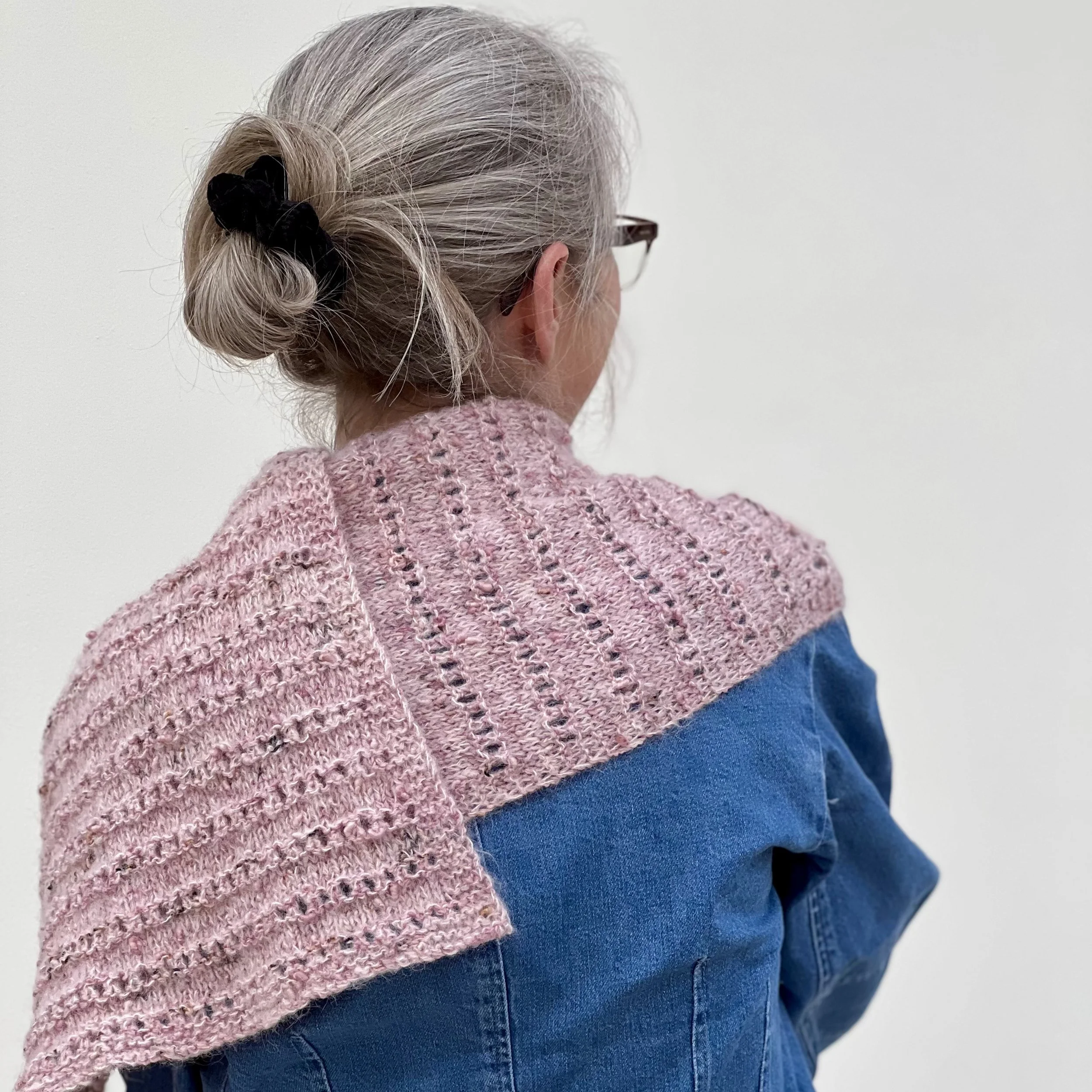 Emma is wearing the large Parallel Lines Shawl drape over her shoulder. She has turned away from the camera to show the shawl's lower border.
