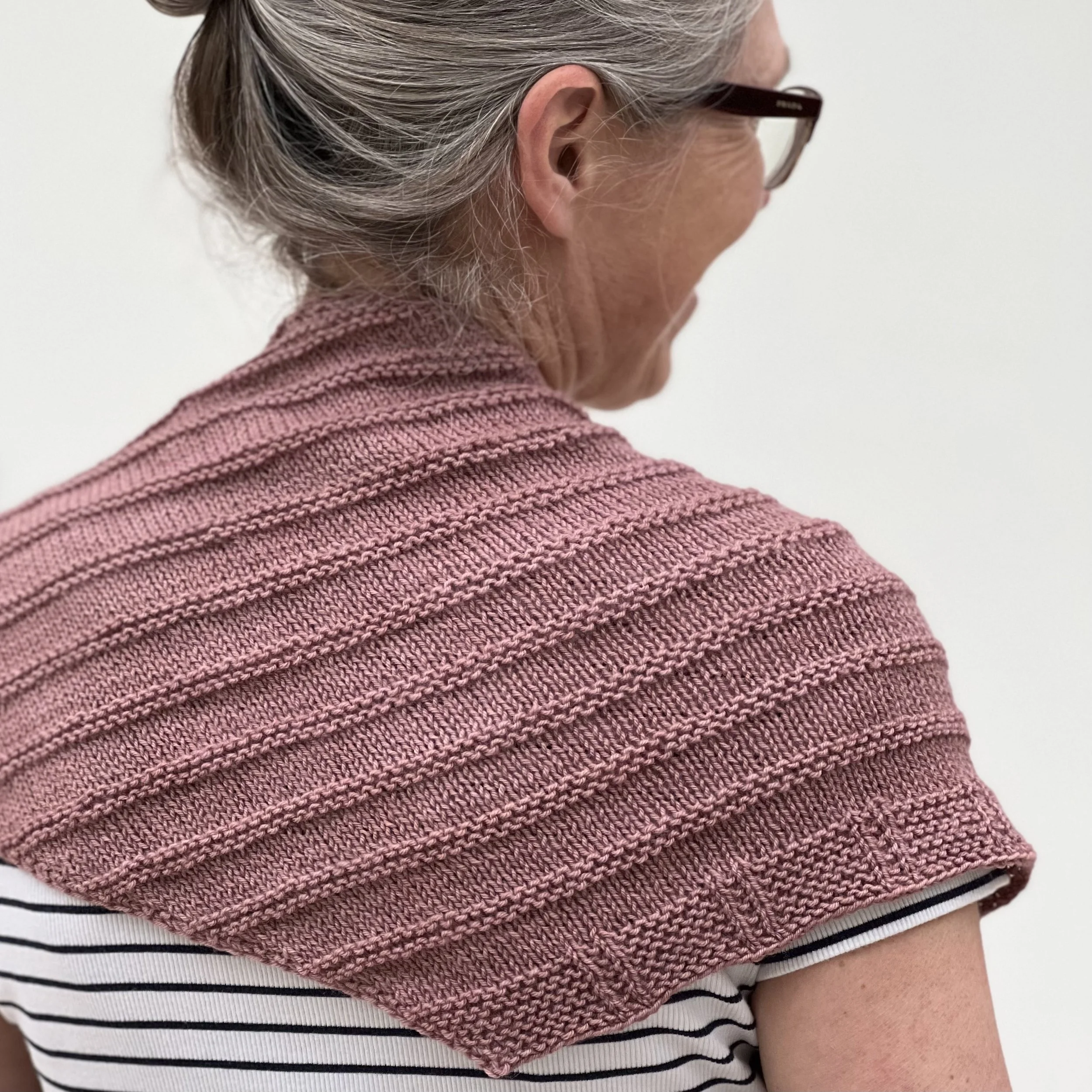Emma is wearing the knitted Lyra Shawl and has turned away from the camera to show the ridged stitch pattern and the shawl border.