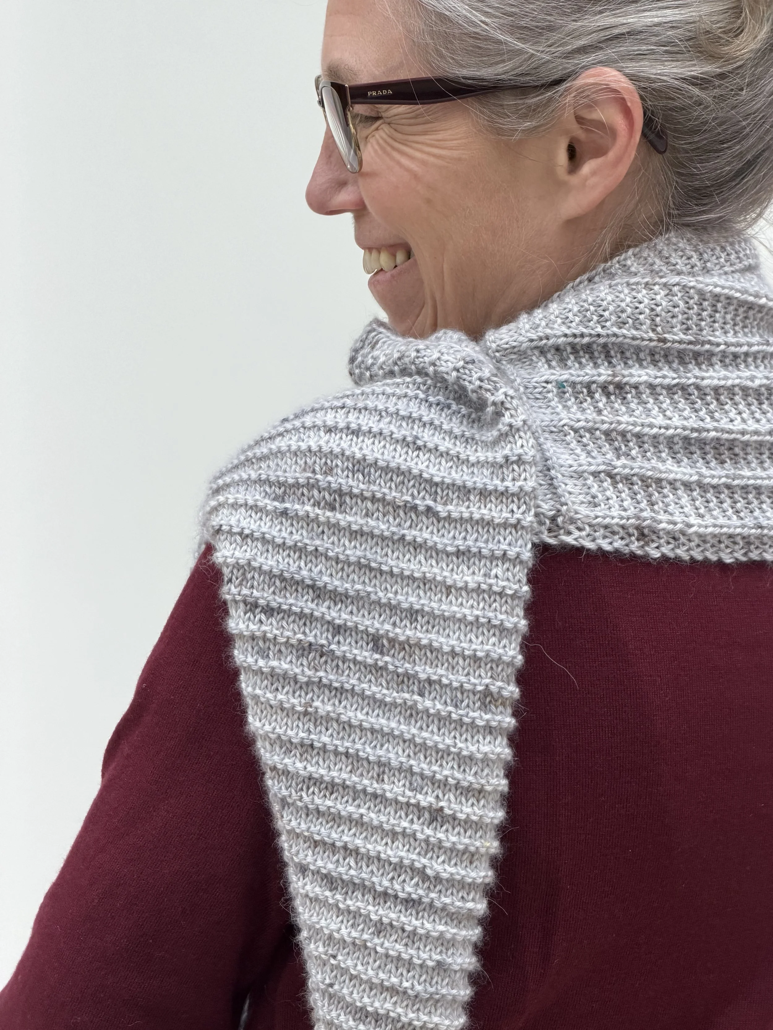 Emma is wearing the knitted Level Crossing Shawl and has turned away from the camera to show the ridged stitch pattern.