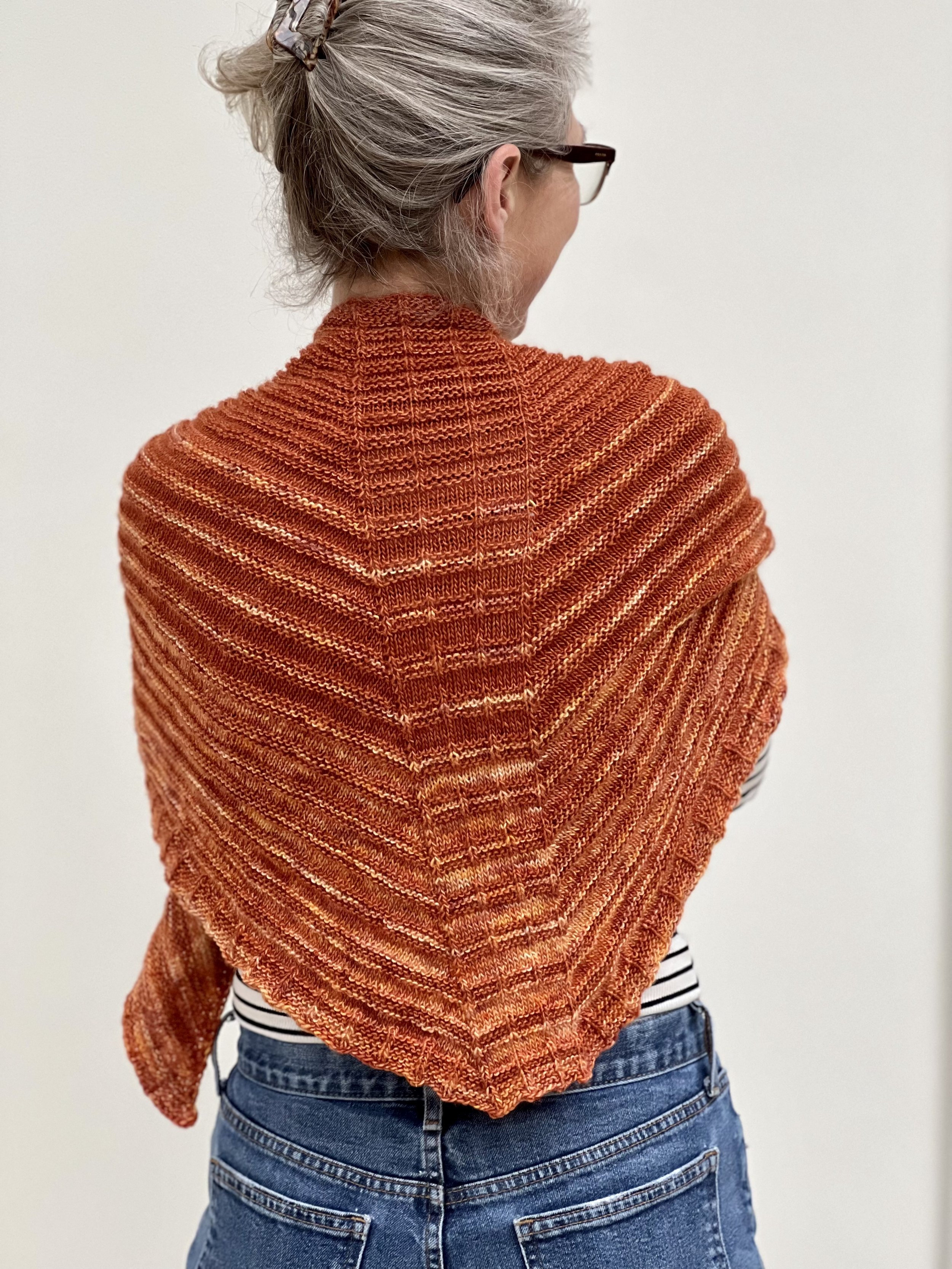 Emma is wearing the Forvie knitted shawl and has turned away from the camera to show the ridge pattern and subtle brown and orange shades.