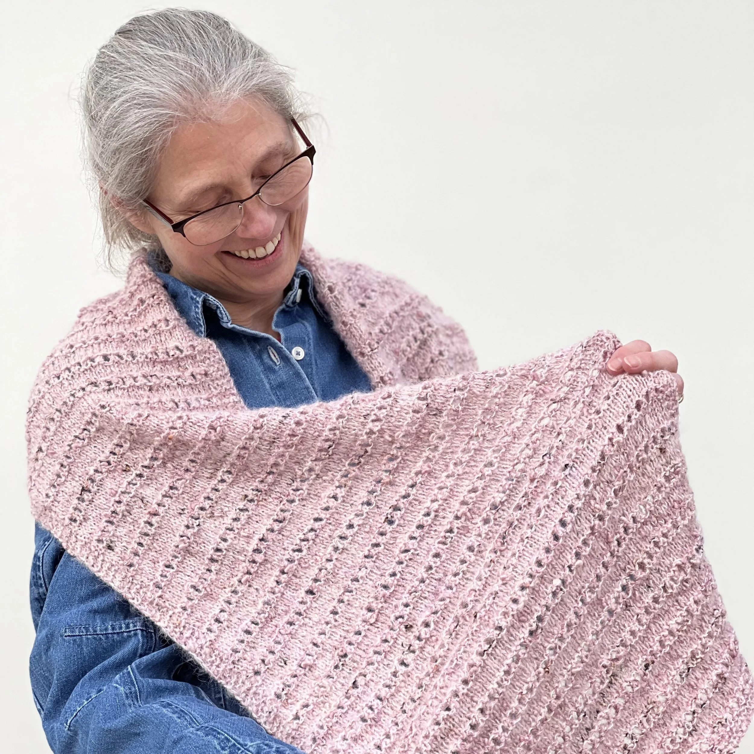 Emma is wearing the large knitted Parallel Lines Shawl and is holding one wide end open to the camera to show the ridged eyelet stitch pattern