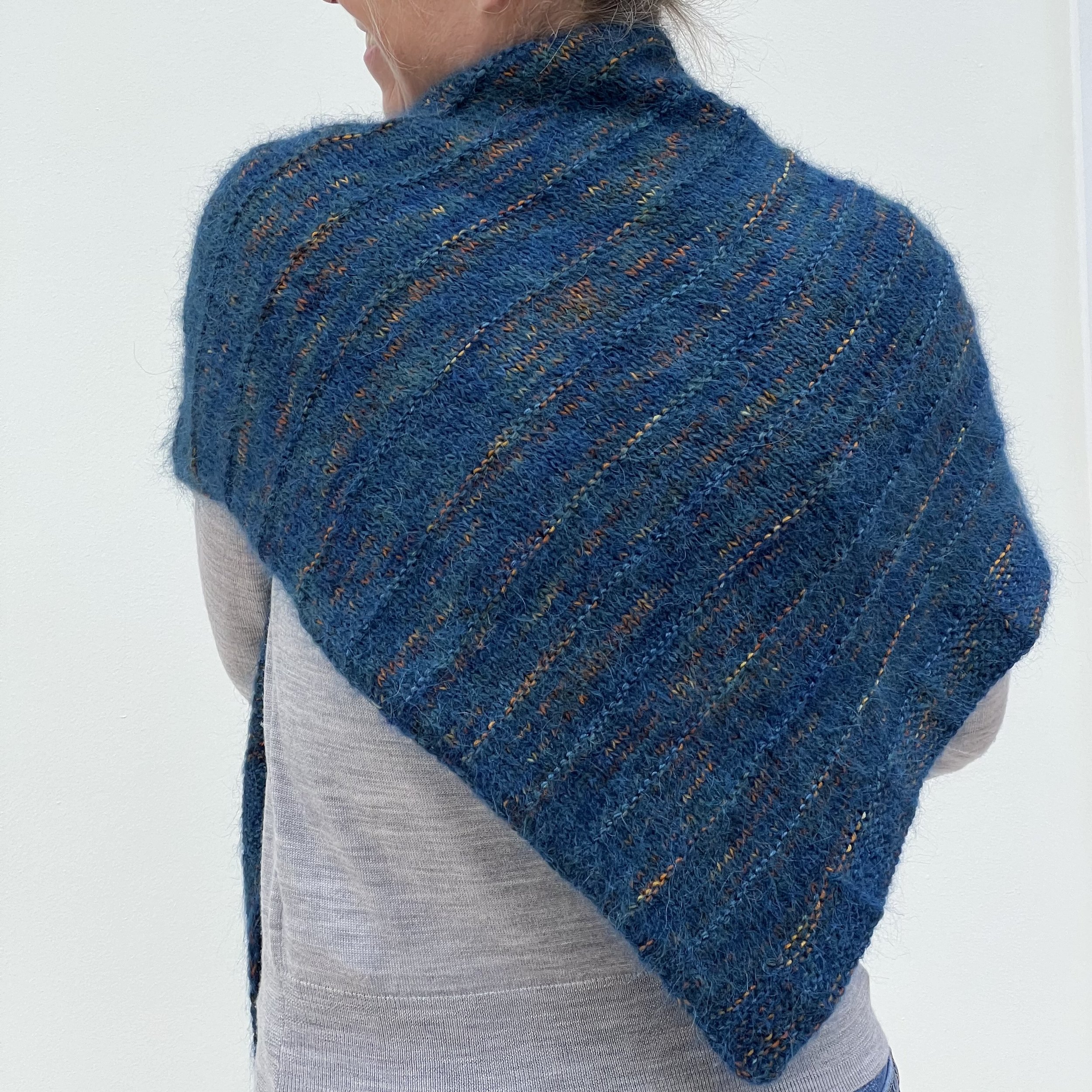 Emma is wearing the Deneb knitted shawl and has turned away from the camera to show the ridge pattern and subtle blue and orange shades.