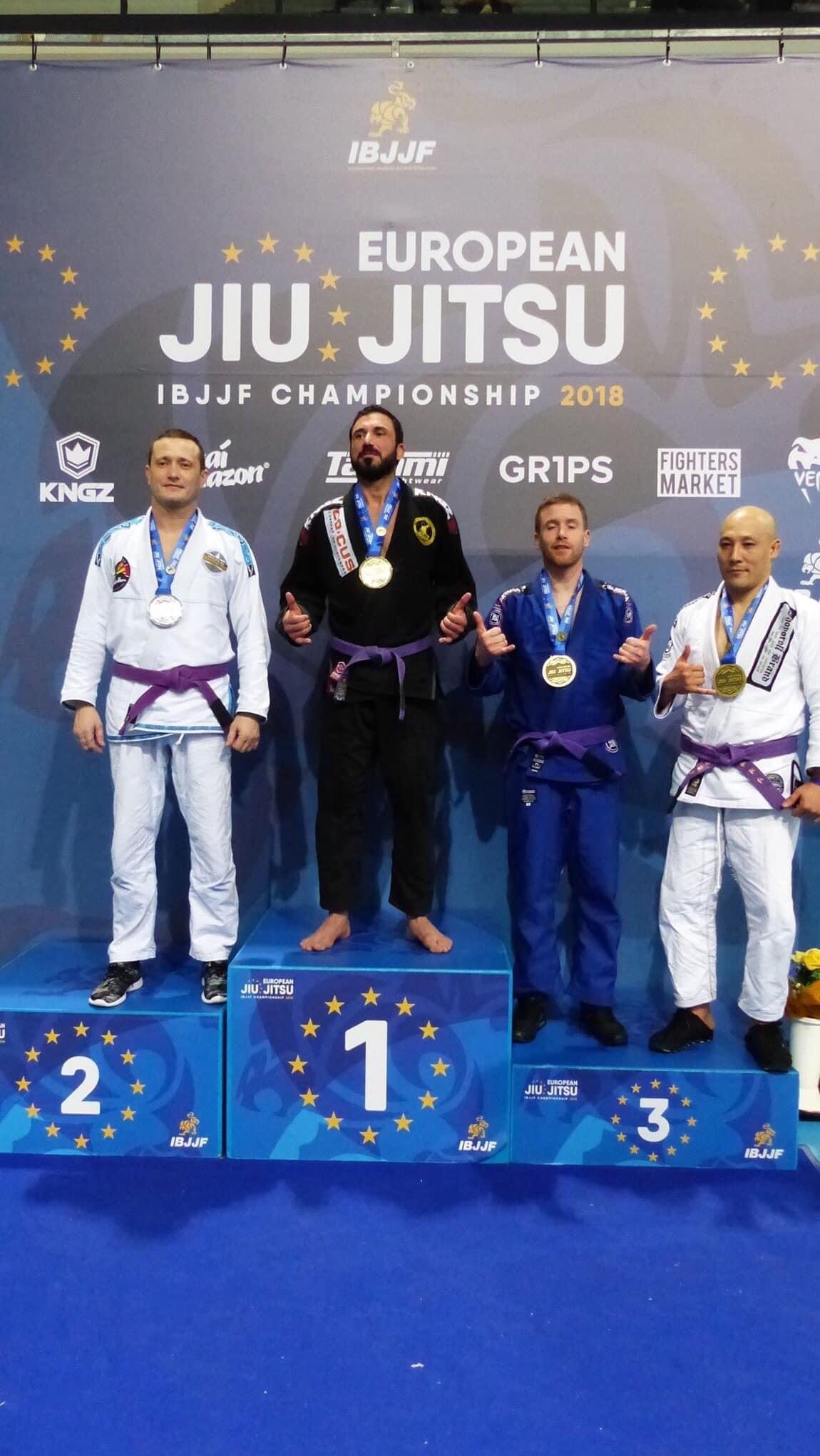 Siegerpodest bei den European Jiu Jitsu Championships 2018 mit vier männlichen Teilnehmern in Jiu Jitsu-Anzügen, Medaillen um den Hals, auf den ersten drei Plätzen, im Hintergrund ein Banner mit der Aufschrift 'European Jiu Jitsu IBJJF Championship 2018'.