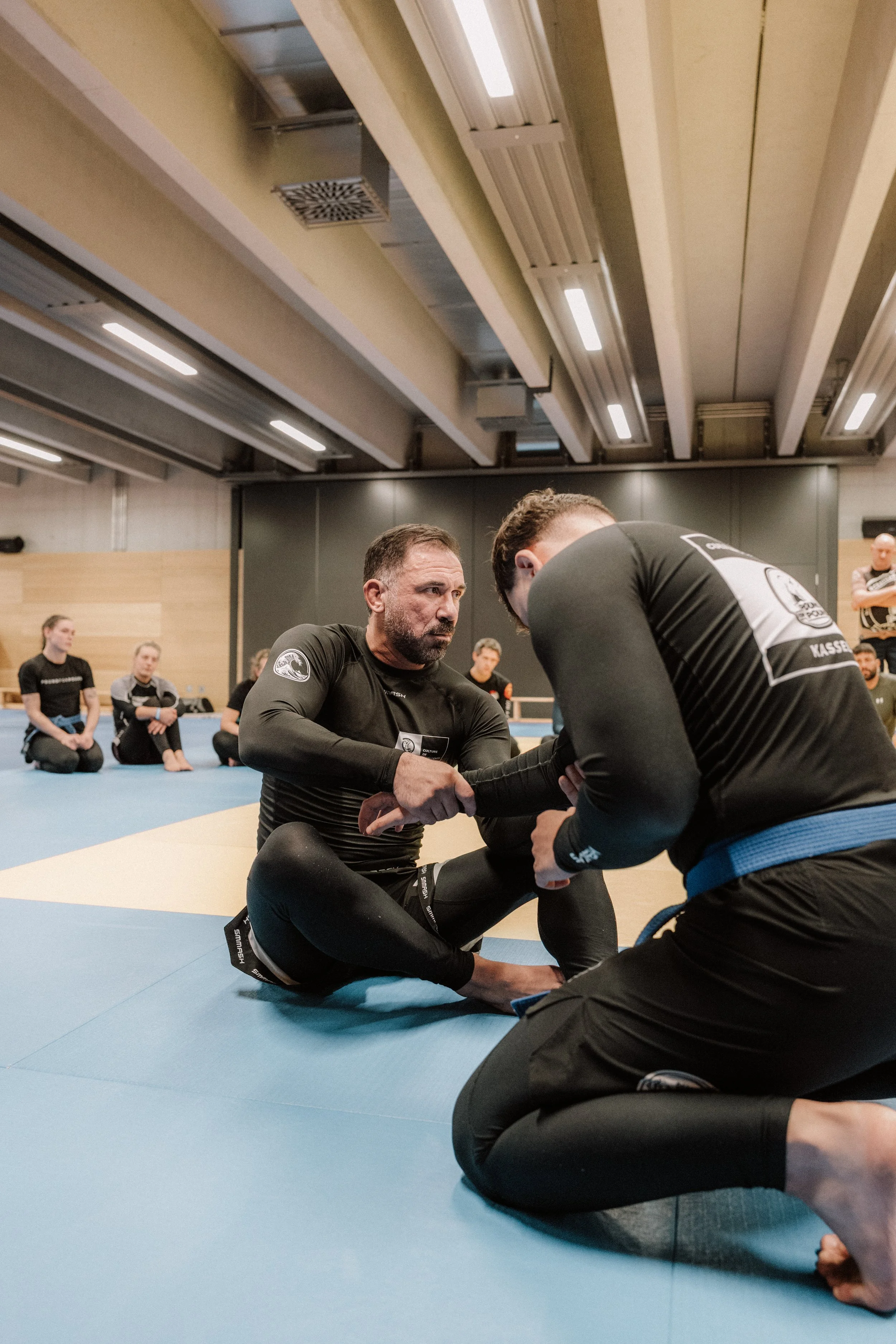 Zwei Männer beim Brazilian Jiu-Jitsu Training, einer zeigt eine Technikorientierte Position, umgeben von anderen Trainierenden im Trainingraum.