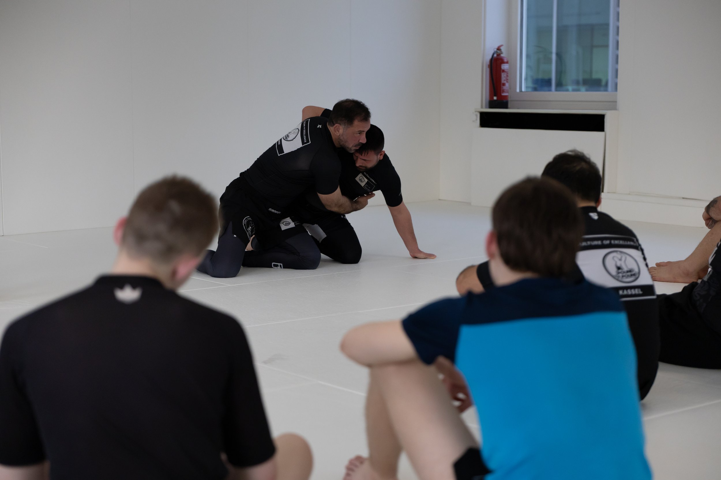 Zwei Trainer zeigen einer Gruppe von Schülern im Kampfsport auf dem Boden Techniken im Mattenraum.