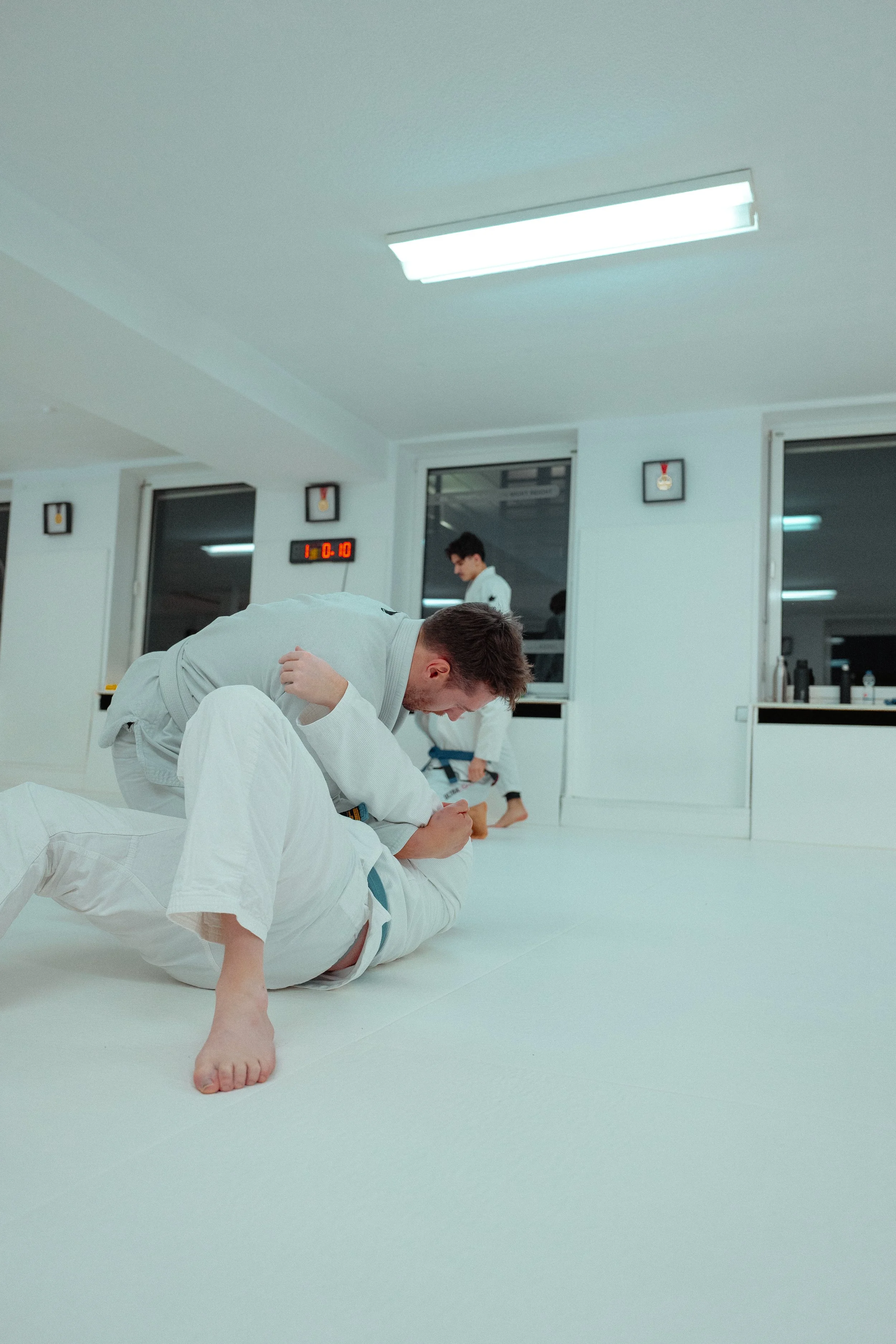 Zwei Judoka in weißen Gi, die eine Grappling-Übung auf der Matte durchführen, in einer Turnhalle mit weißem Boden.