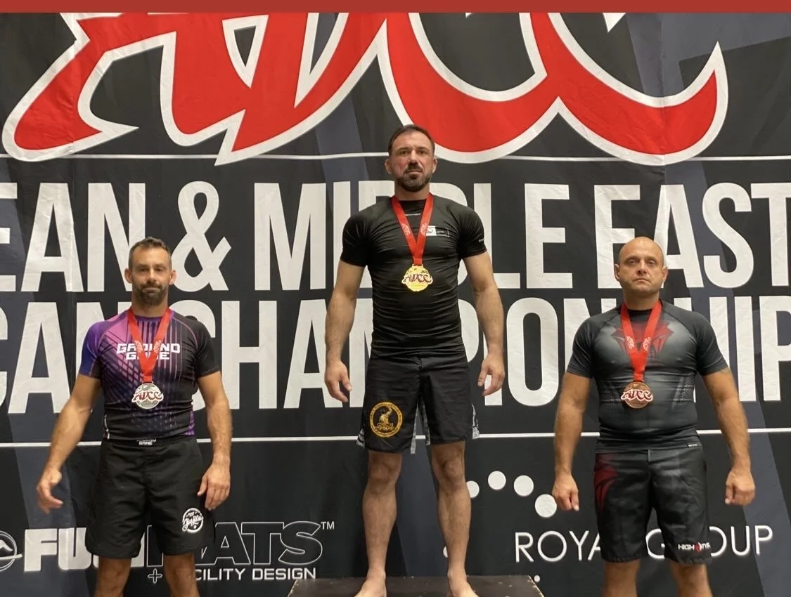 Drei Männer auf dem Siegerpodest bei einer Strongman-Meisterschaft, tragen Medaillen um den Hals und stehen vor einem schwarzen Hintergrund mit weißen Schriftzügen.