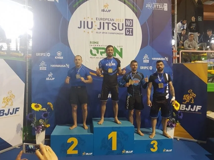 Vier Männer auf dem Siegerpodest bei einem Jiu Jitsu Wettkampf, mit Medaillen um den Hals, auf einer blauen Bühne mit Logos und Texten im Hintergrund. Die ersten drei Plätze sind mit Nummern 1, 2, 3 gekennzeichnet.