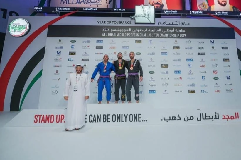 Drei Männer in Jiu-Jitsu-Anzügen mit Medaillen stehen auf dem Siegerpodest bei einem Turnier in Abu Dhabi, ein weiterer Mann in traditioneller arabischer Kleidung steht davor.