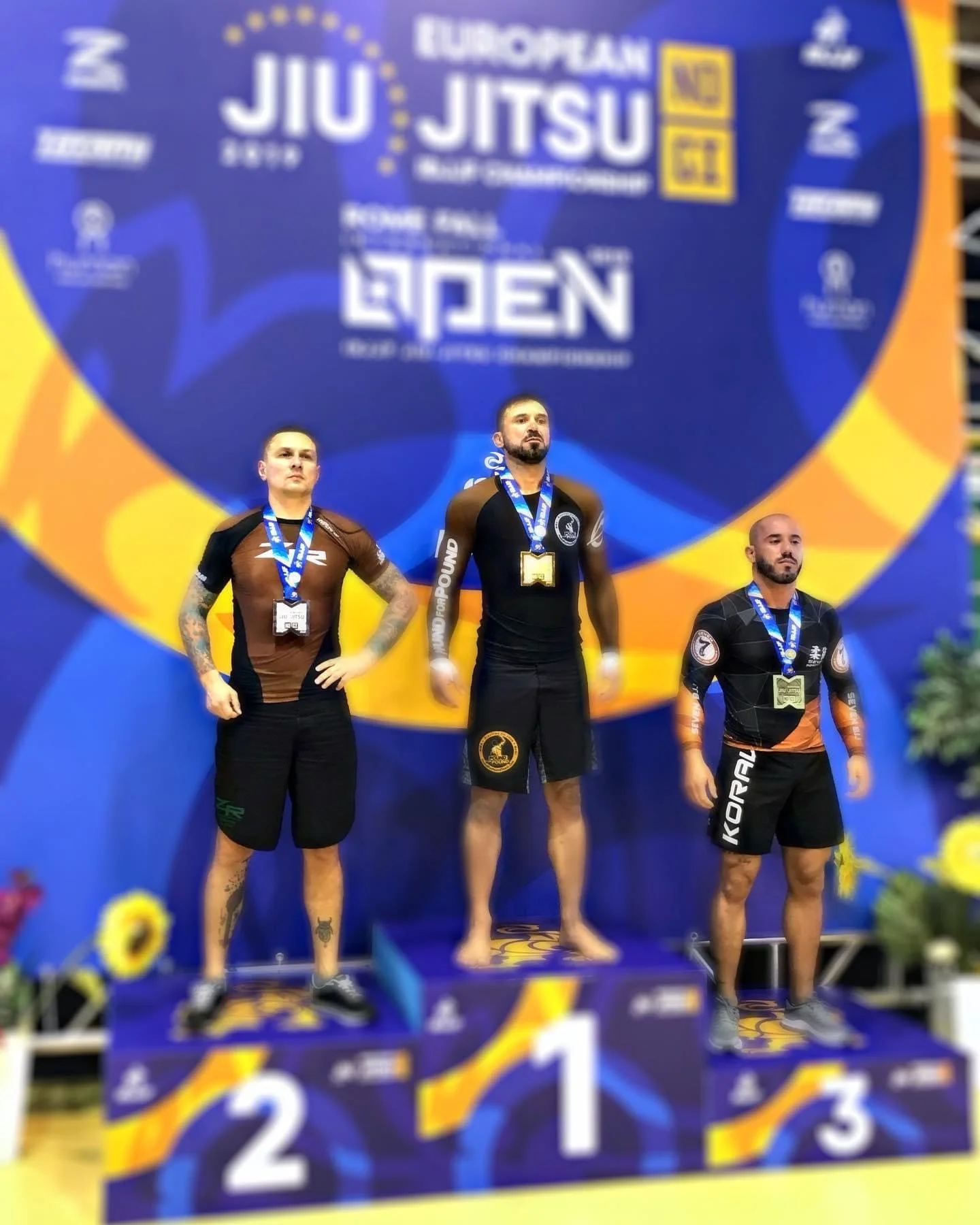 Drei Männer stehen auf einer Siegerpodest während eines Jiu-Jitsu-Wettbewerbs, mit Medaillen um den Hals, vor einem großen blauen Banner mit dem Text "European Jiu Jitsu".