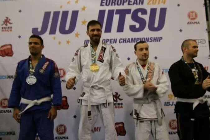 Vier Judoka auf dem Podium bei einer Jiu-Jitsu-Meisterschaft, der mittlere trägt eine goldene Medaille, die anderen Silber und Bronze, im Hintergrund ein Banner mit 'EUROPEAN 2014 JIU JITSU'.