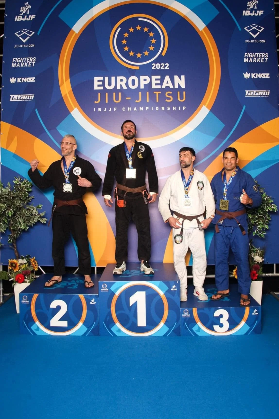 Siegerpodest bei der europäischen Jiu-Jitsu-Meisterschaft 2022 mit vier Kämpfern in Gi-Uniformen, Medaillen und Glückwünschen, im Hintergrund eine große blau-orange Gestaltung mit Veranstaltungstitel und Logos.