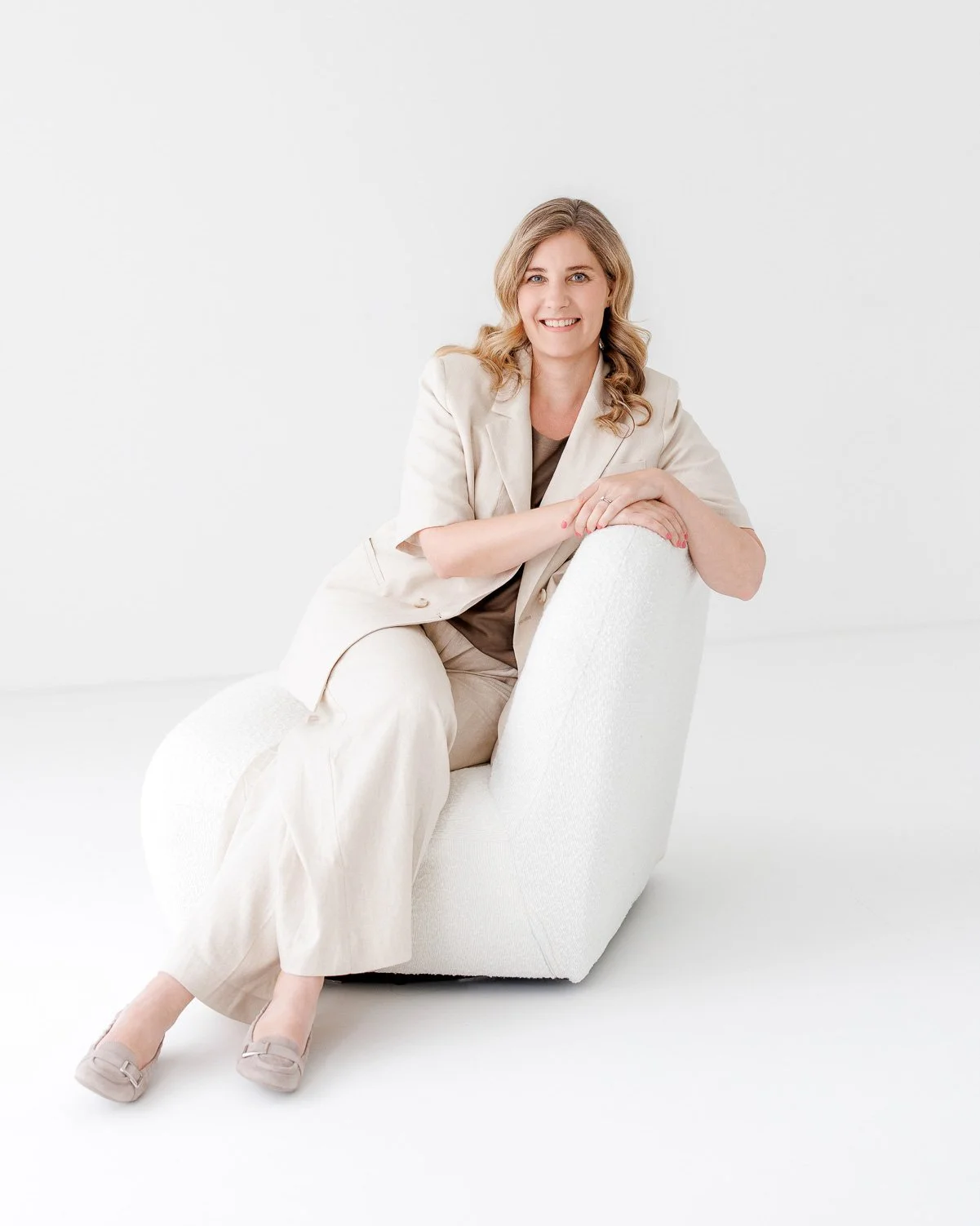 A woman with wavy blonde hair, wearing a beige blazer and pants, sitting on a white plush chair in a minimalist white room. She is smiling and looking at the camera.