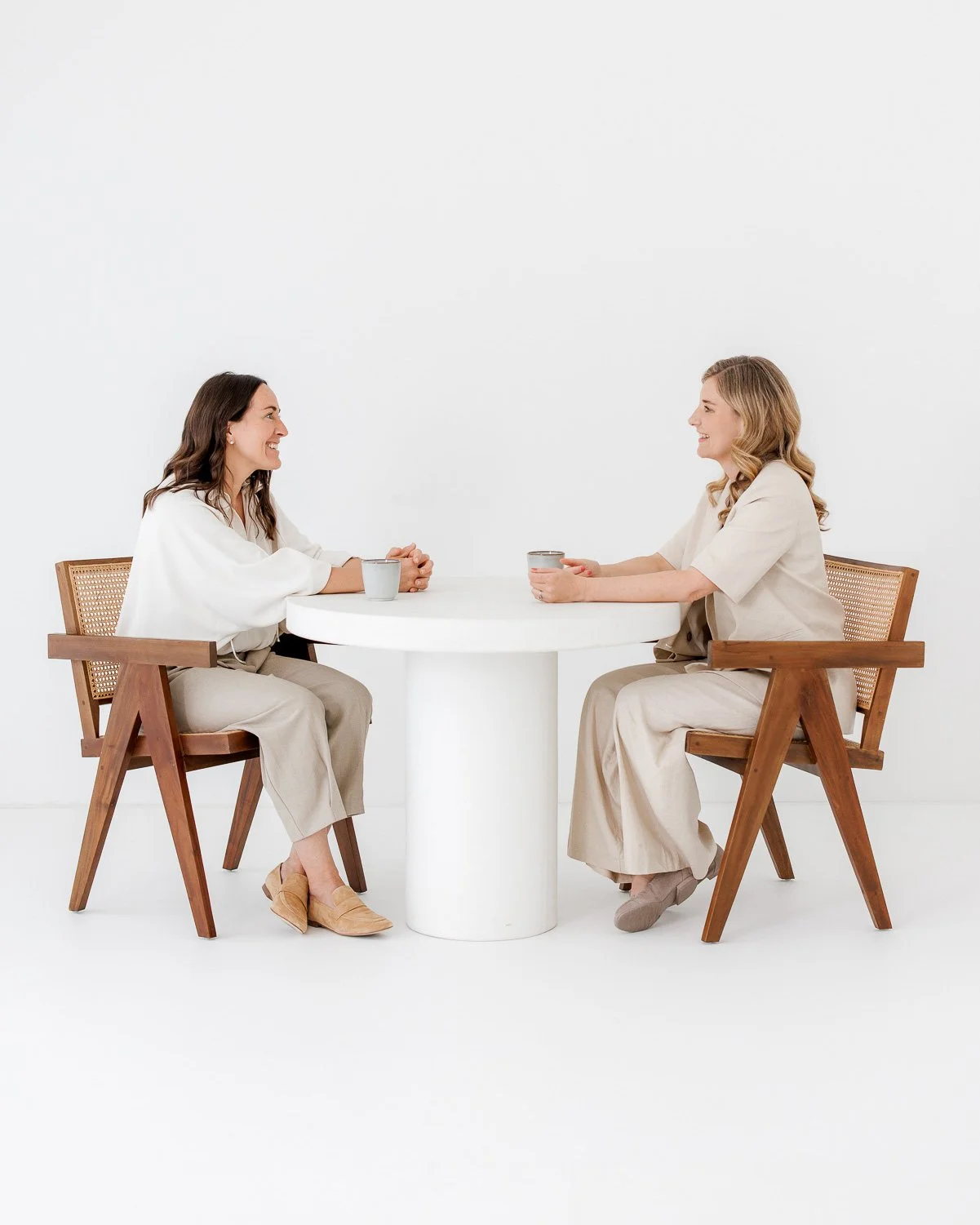 Two women sitting and talking at a white round table, each holding a cup, in a minimalistic white room.