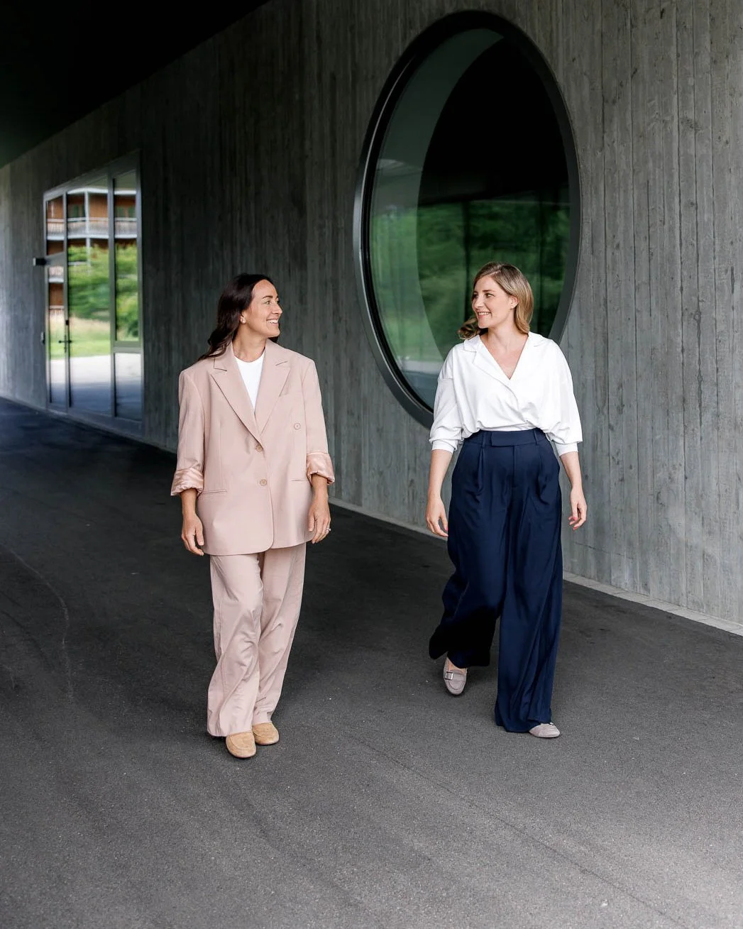 Two women walking and talking outside a modern building with a large round window, dressed in professional attire.