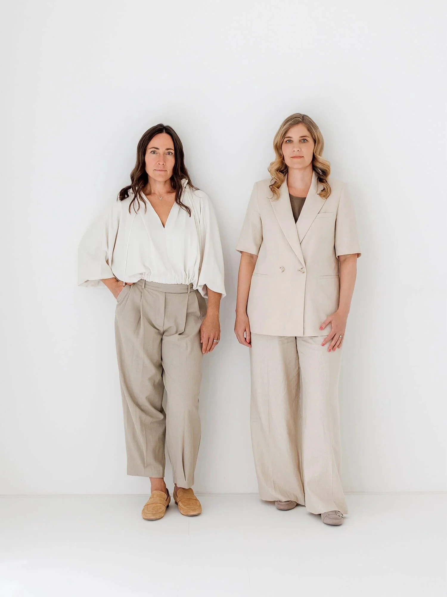 Two women stand against a white wall, dressed in neutral-colored professional outfits, facing forward with serious expressions.