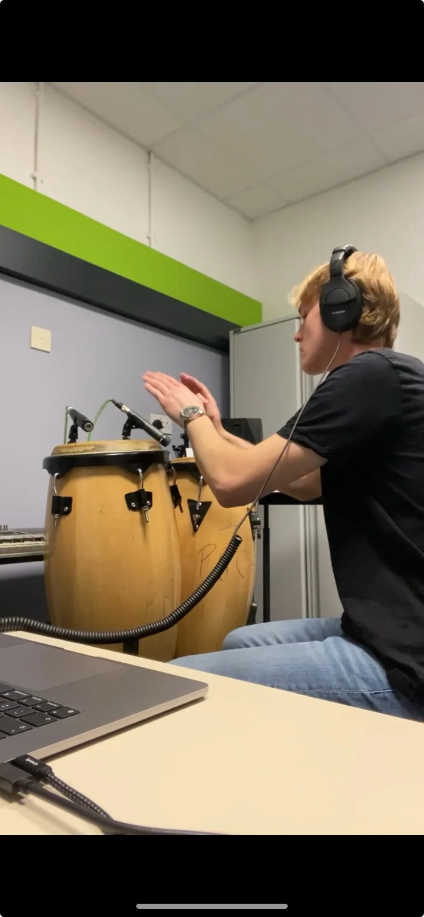 A person wearing headphones playing conga drums in a recording studio.