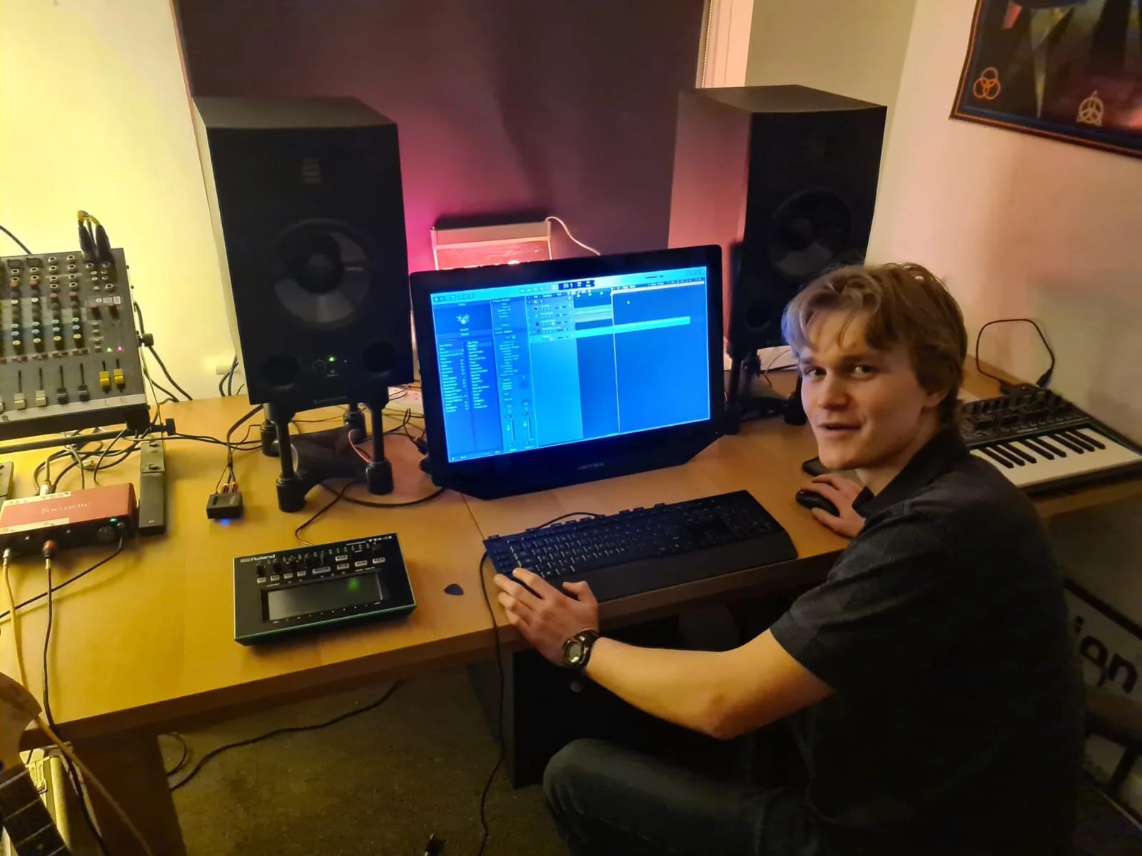 A young man sitting at a desk in a home music studio, using a computer with a digital audio workstation software on the screen. The desk has audio equipment, monitors, a MIDI keyboard, and audio cables, with a poster on the wall.