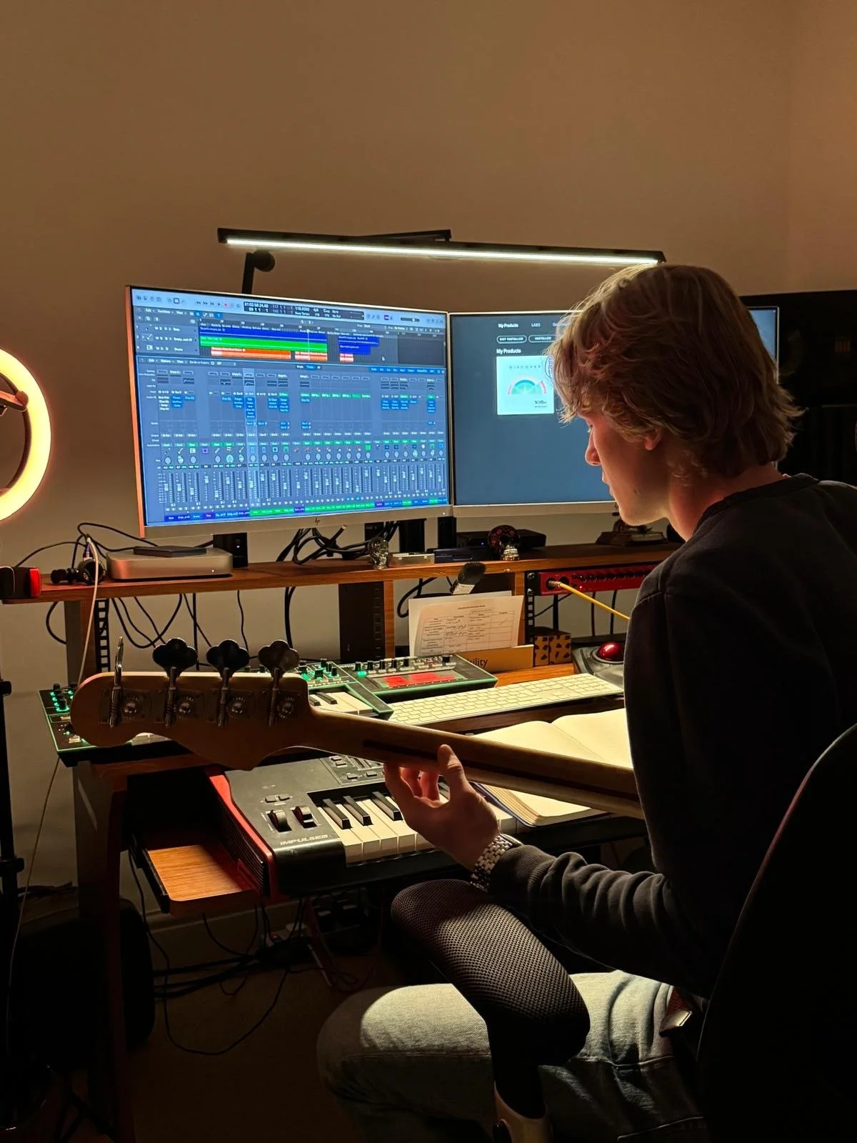 A person with long hair sitting at a desk playing a bass guitar, with two large monitors displaying music production software behind them, and various electronic music equipment on the desk.